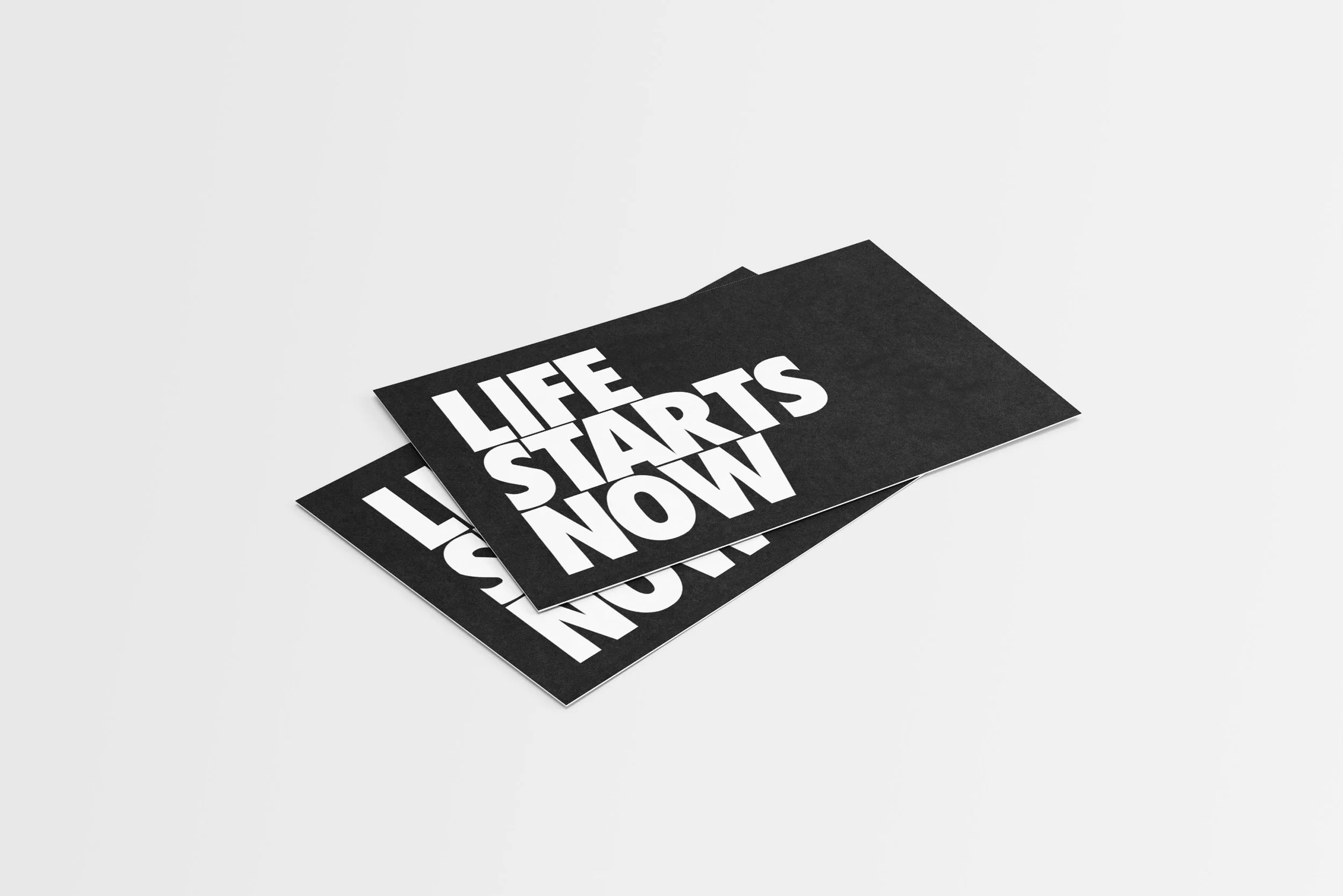 Black paper cards with white bold text reading 'LIFE STARTS NOW,' stacked slightly offset on a white surface.