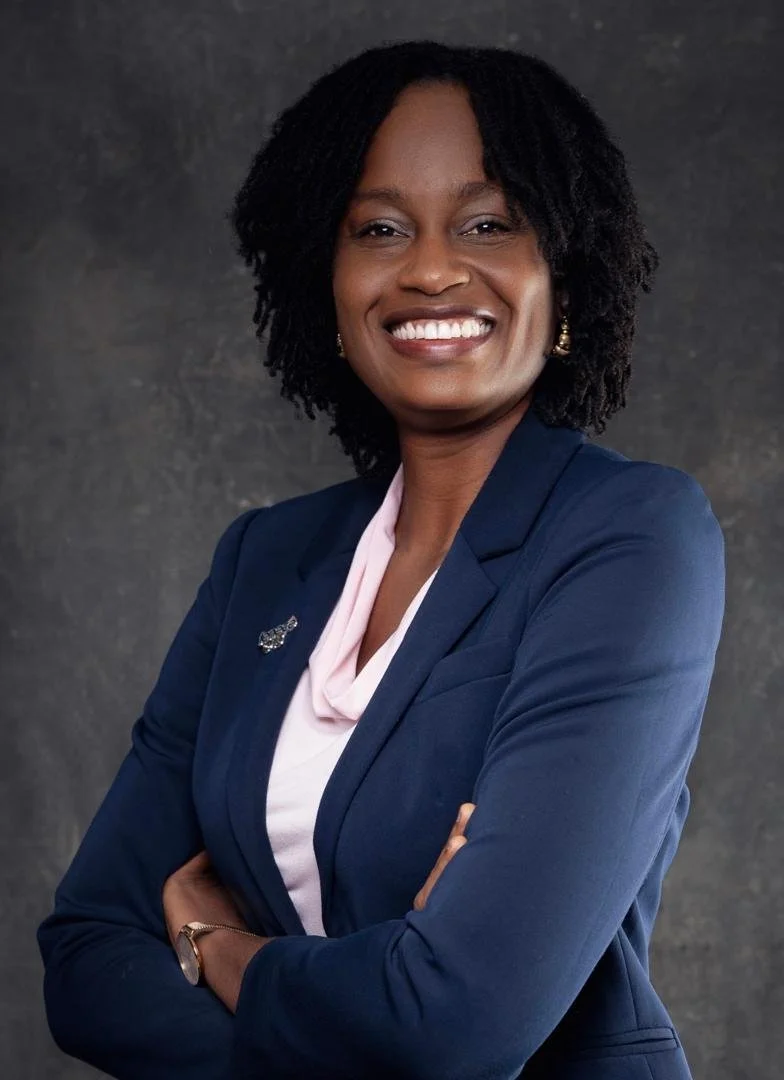 A professional woman with shoulder-length curly black hair, smiling confidently with her arms crossed, wearing a navy blazer, a light pink blouse, and gold earrings, against a dark textured background.