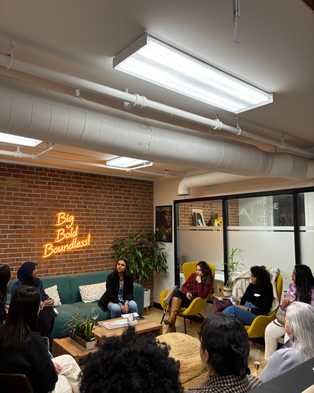 Women in a meeting or discussion in a modern office lounge with brick walls, a neon sign that reads 'Big Bold Boundless!', and various plants.
