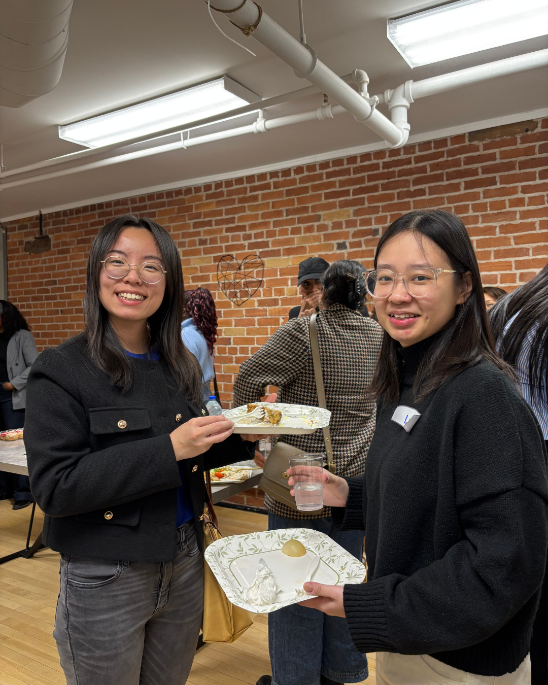 Two young women smiling and holding plates with food at a social gathering in a room with a brick wall.
