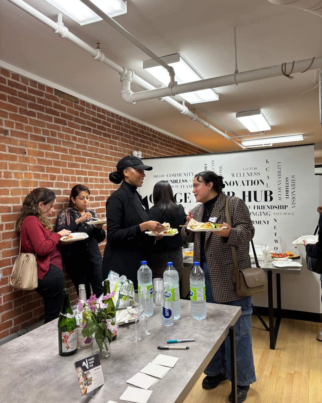 People standing in line at a buffet table, serving themselves food at an indoor event with a brick wall background and informational wall art.