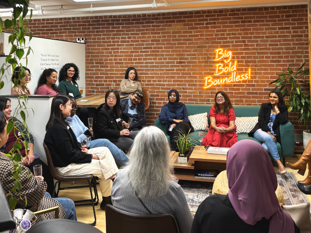 A diverse group of women in a cozy, modern room with a brick wall, sitting and listening at a seminar or panel discussion. Some hold glasses of wine; three women are seated on a teal couch, one is standing behind a wooden counter. A neon sign on the wall reads 'Big Bold Boundless!'. There is a plant on the coffee table and a whiteboard with text in the background.