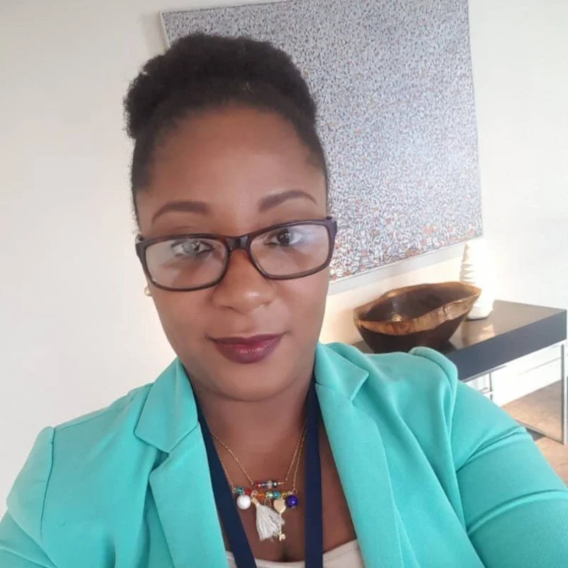 A woman with glasses and styled hair taking a selfie indoors, wearing a light blue blazer and a colorful necklace, with a decorative wooden bowl and artwork on the wall in the background.