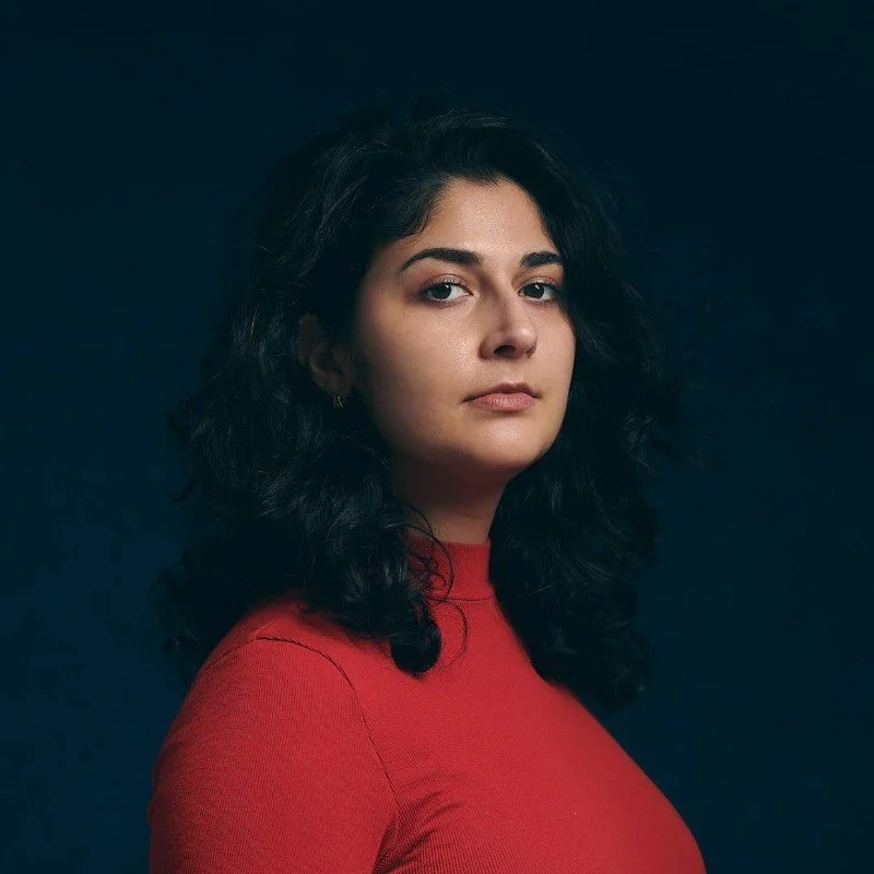 A woman with dark, curly hair wearing a red top looks at the camera against a dark background.