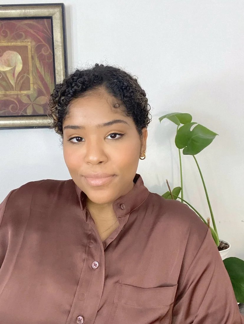A young woman with short curly hair and gold earring, wearing a brown satin button-up shirt, sitting indoors near a green potted plant with large leaves, and a framed artwork on the wall.