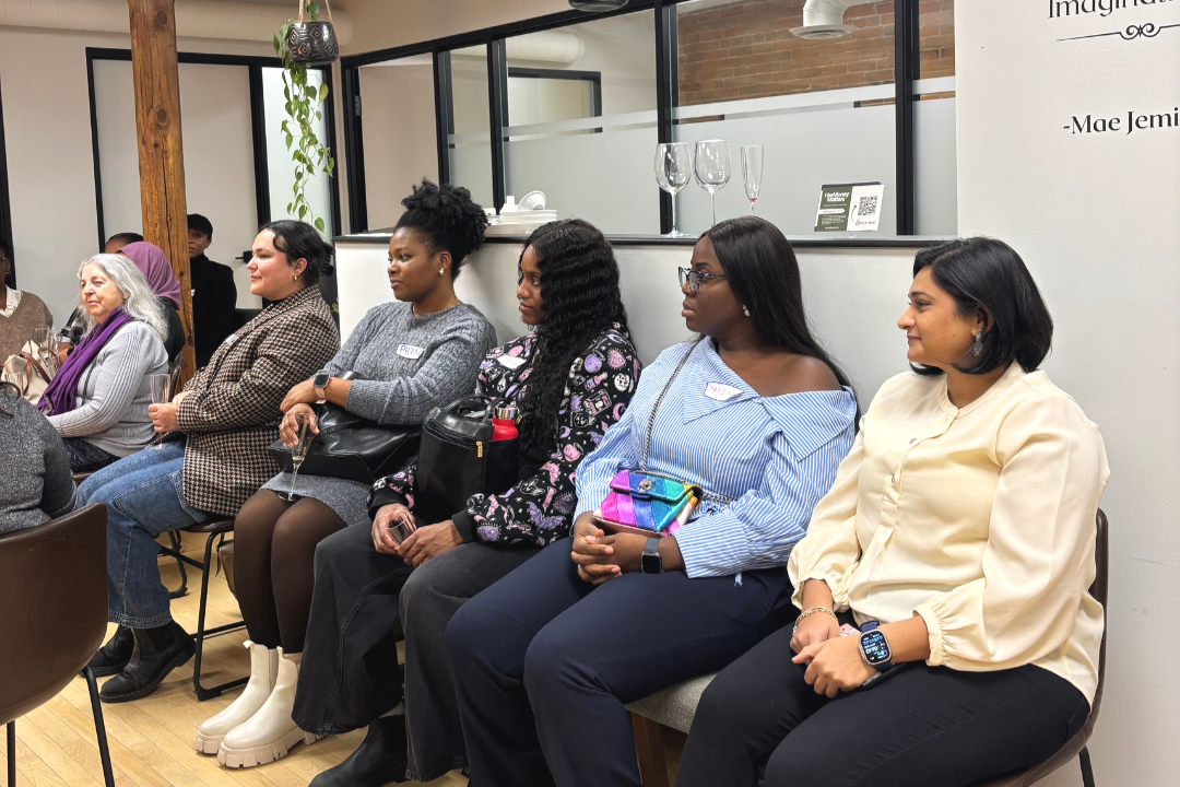 Side view of a group of women sitting in a row at an indoor event, attentively listening. The background shows a modern, stylish interior with a glass partition, decorative plants, and wine glasses displayed on a shelf.