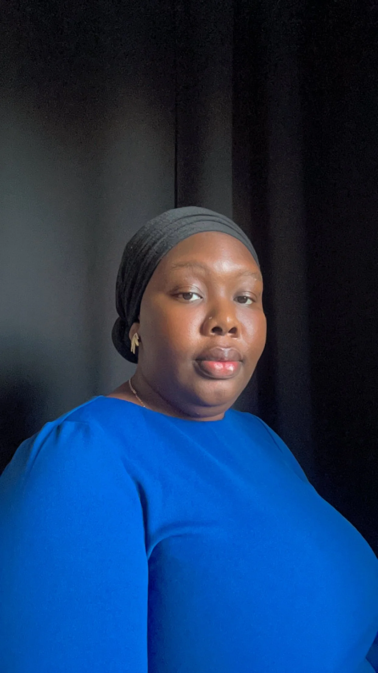 A woman with dark skin wearing a black headscarf and a blue top, seated against a black background.