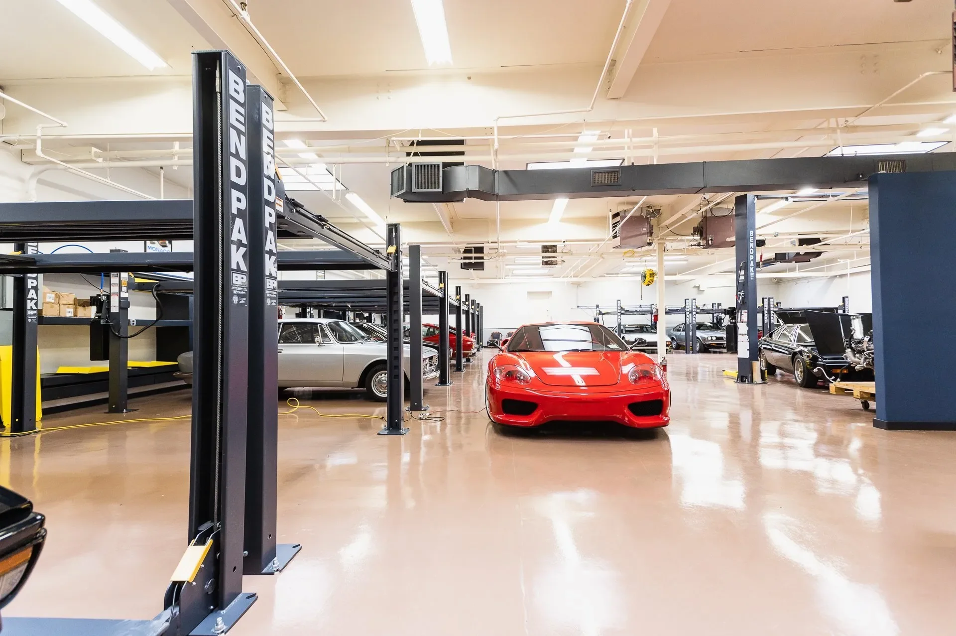 An indoor car storage or showroom with multiple classic and sports cars, including a prominent red sports car at the center, surrounded by various other vintage and luxury vehicles.