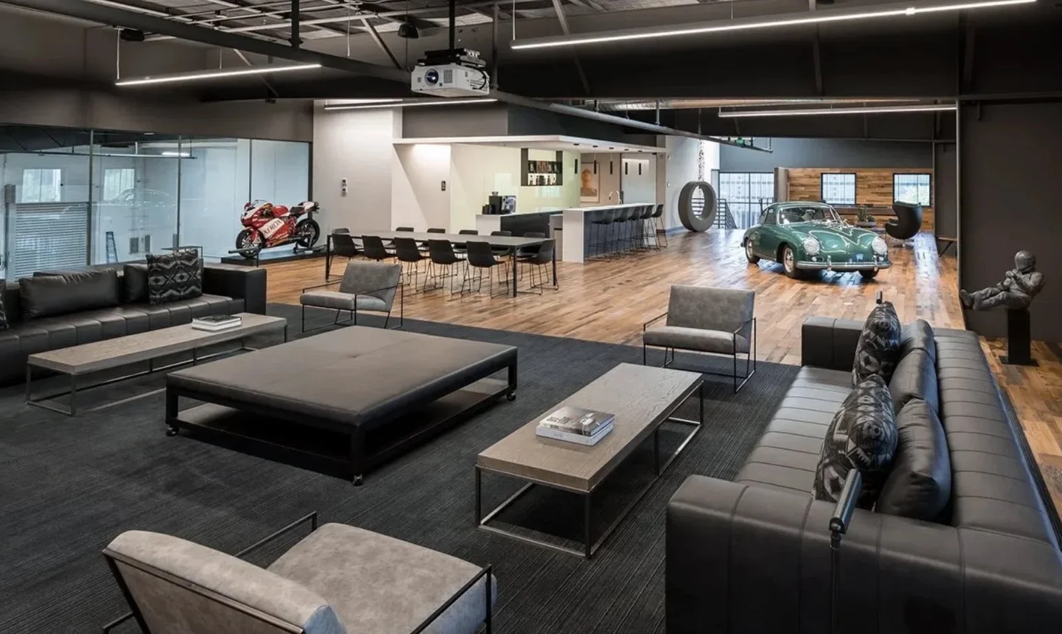Modern industrial-style lounge with black, gray, and beige furniture, a large black rug, and a wooden floor. In the background, a vintage green sports car, a red and white motorcycle, and a sculpture of a person sitting on a bench are visible. There is a bar area with black chairs, a large circular sculpture, and a glass partition.