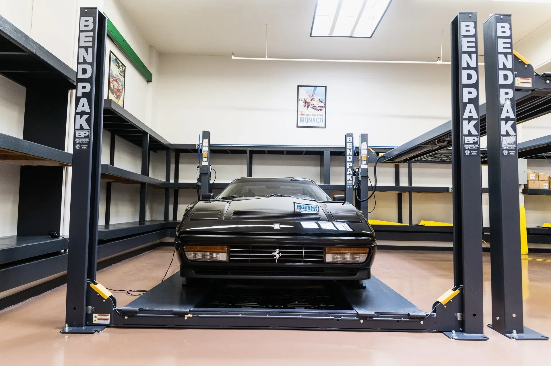 A black Ferrari car on a BENDPAK car lift in a room with shelves and posters on the wall.