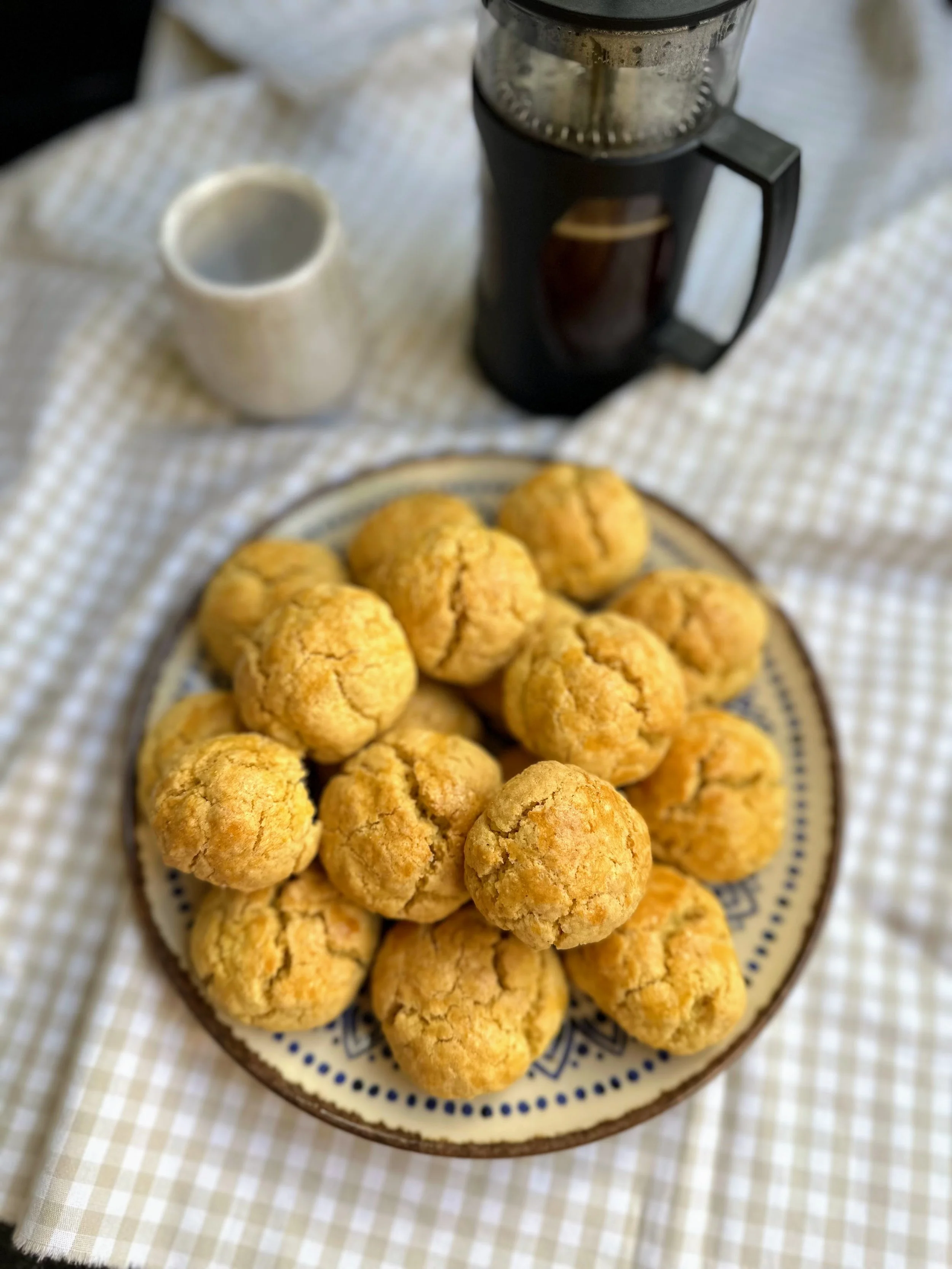Cookies de milho em um prato decorativo sobre uma toalha xadrez, com uma cafeteira francesa ao fundo.
