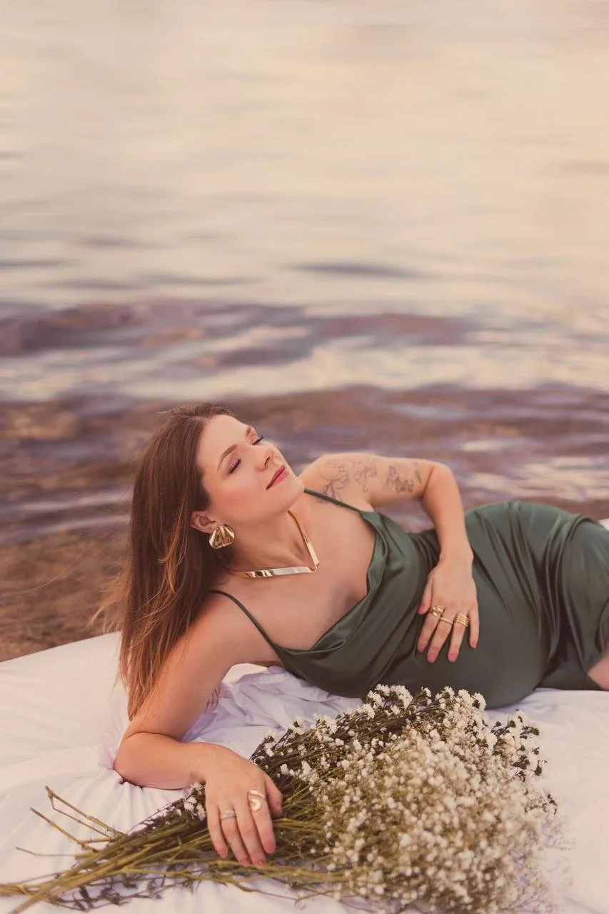 Mulher de vestido verde deitada em uma cama ao lado de um rio, segurando um buquê de flores brancas, com tatuagens no braço e acessórios dourados.