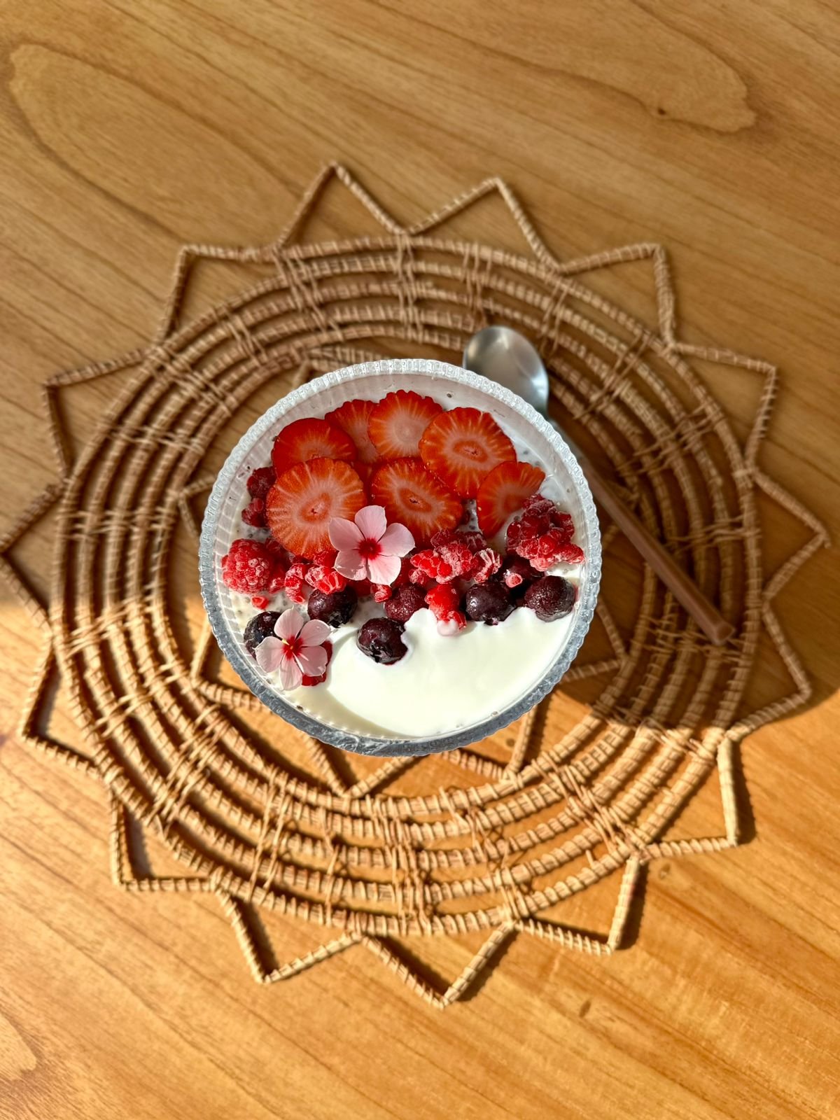 Taça de sobremesa com morangos fatiados, frutas vermelhas, flores comestíveis e creme, sobre uma base de palha de trigo, colocada sobre uma mesa de madeira.