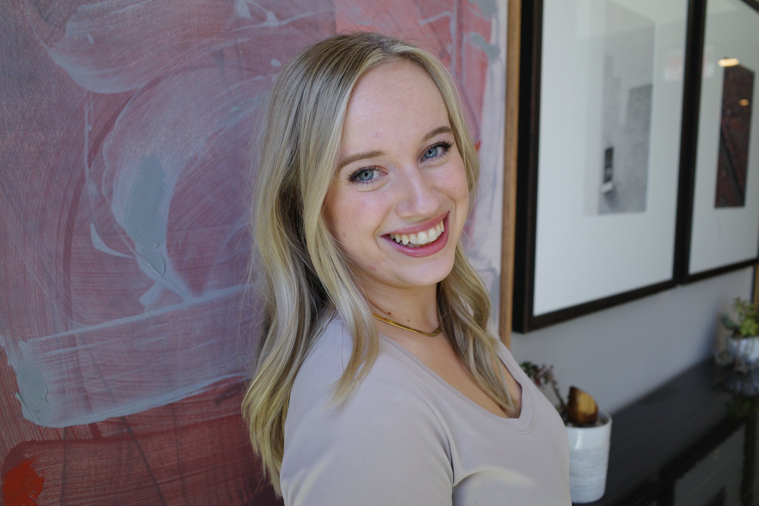 A smiling young woman with blonde hair and blue eyes standing indoors against an abstract pink and gray background.
