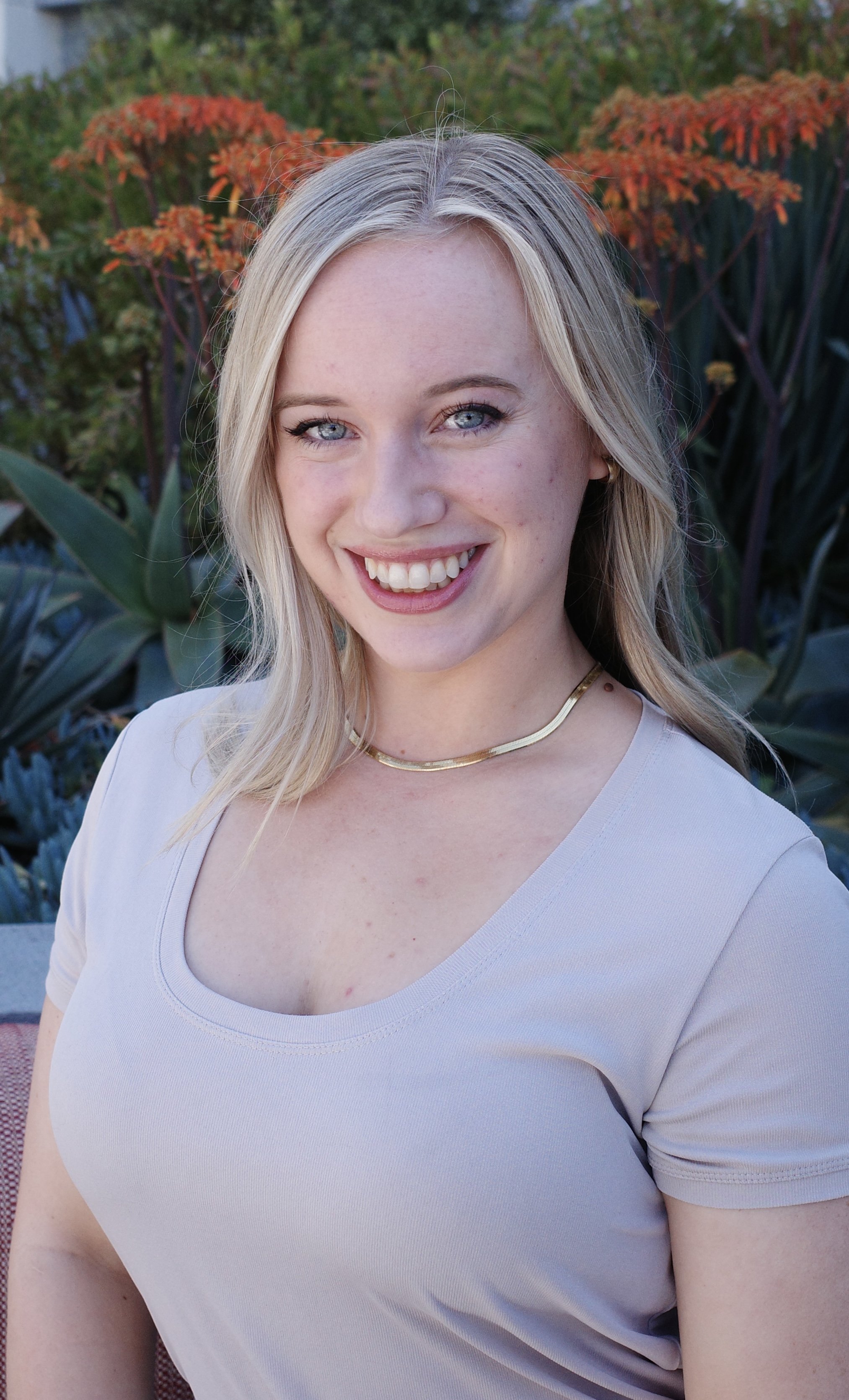 A young woman with blonde hair and blue eyes smiling outdoors, wearing a light-colored top and a gold necklace, with orange and green plants in the background.