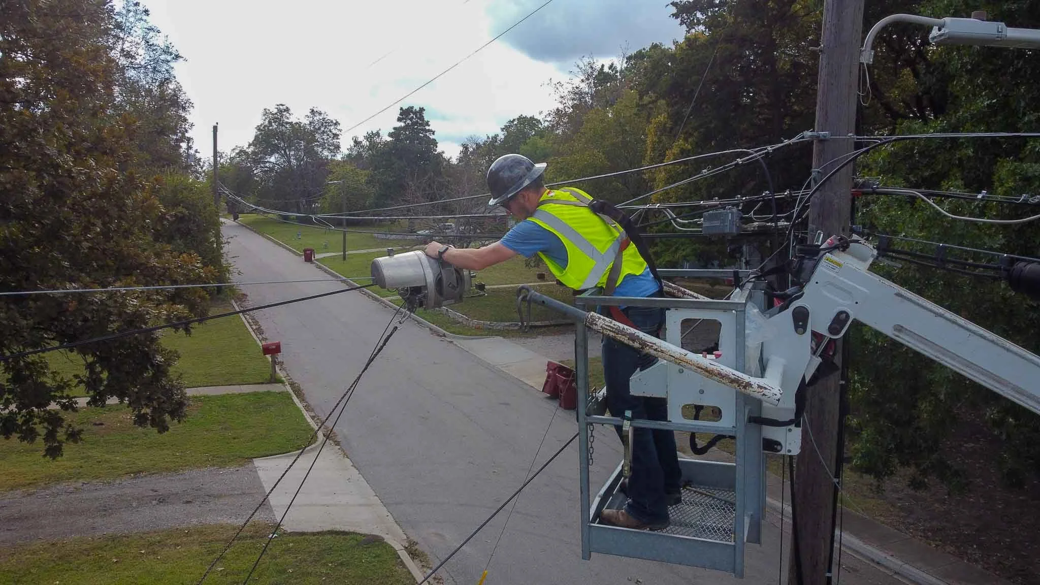 A lineman wearing a hard hat and a high-visibility vest working on power lines from an aerial lift basket beside a wooden utility pole in a residential neighborhood.