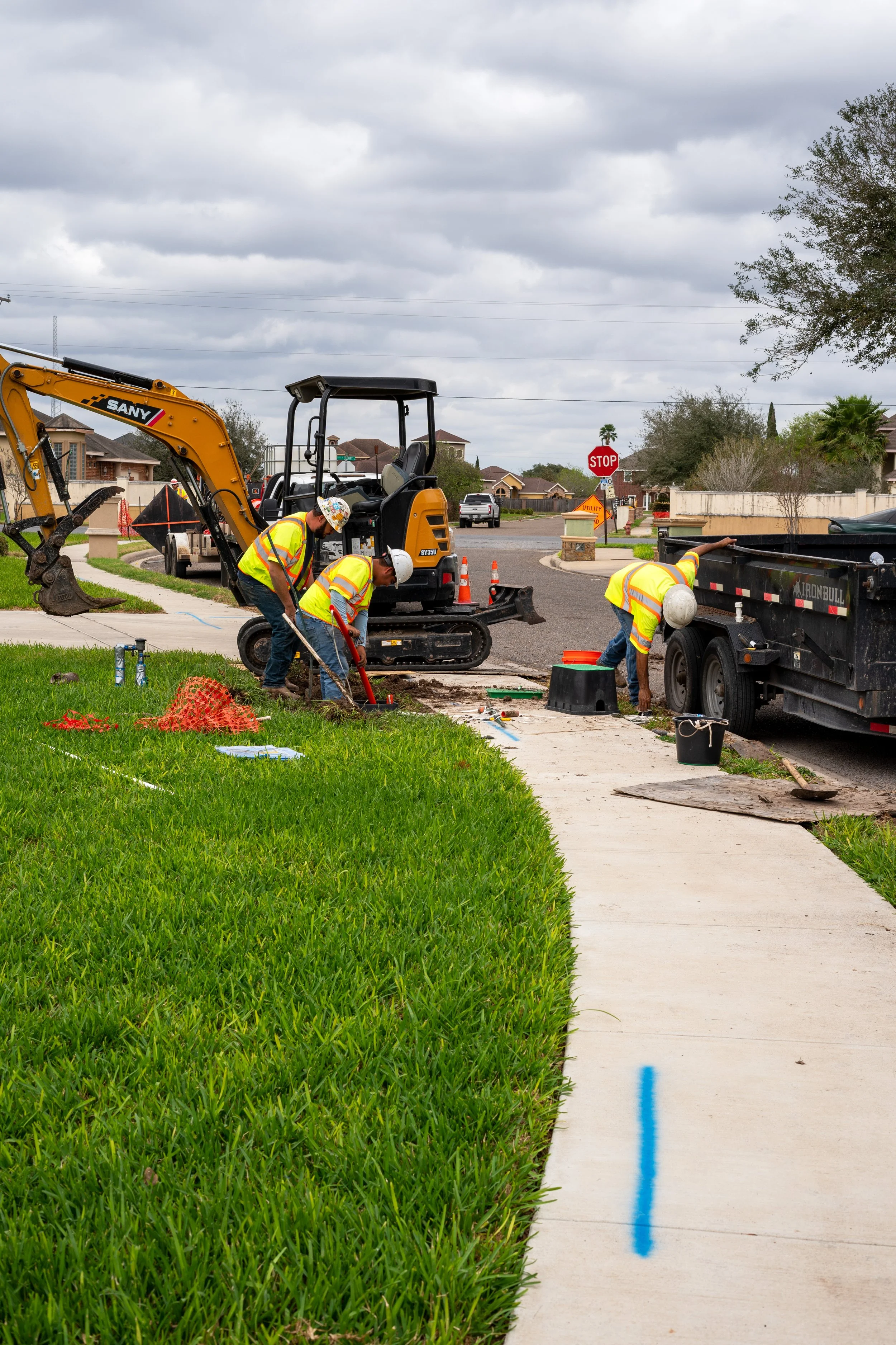 Construction workers wearing yellow vests and hard hats repairing a sidewalk on a residential street, with construction equipment and orange safety cones present.