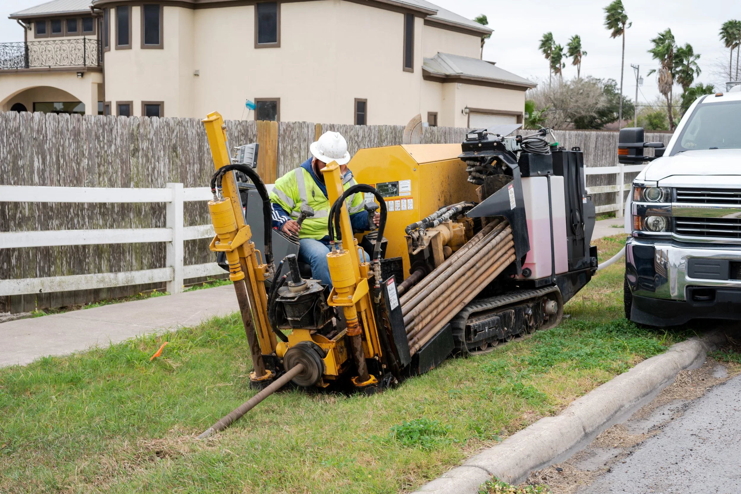 Construction worker in a yellow safety vest and white hard hat operating a large drilling machine next to a parked white truck on a grassy roadside, with a residential area and palm trees in the background.