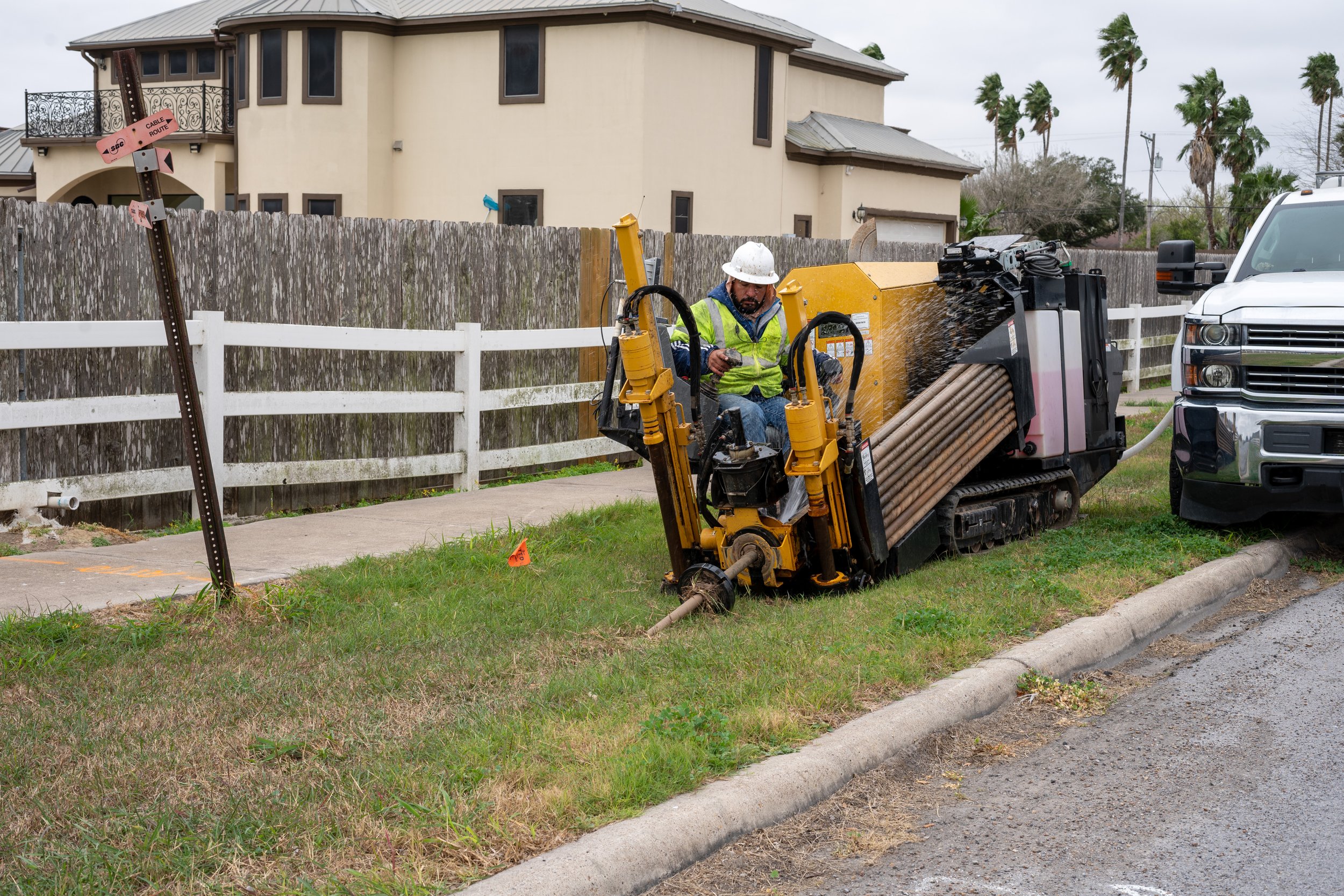 Worker in a yellow safety vest and white hard hat operating a trenchless pipe bursting machine on a grassy lawn next to a concrete curb and a parked white truck.