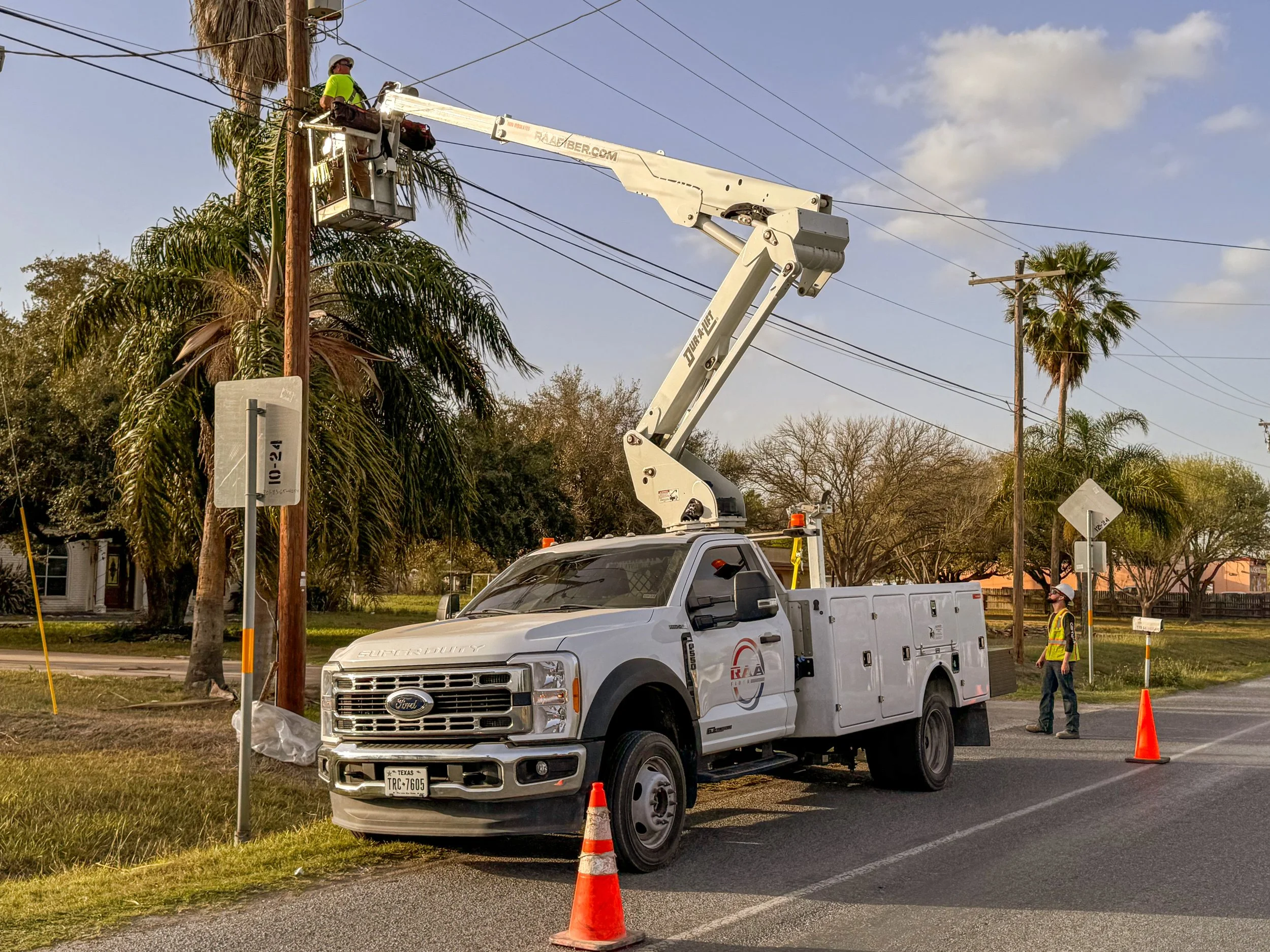 Utility workers repair or maintain power lines using a bucket truck with an extendable arm. One worker is elevated in a bucket checking the overhead wires, while another worker stands on the ground near orange safety cones, observing and supervising.