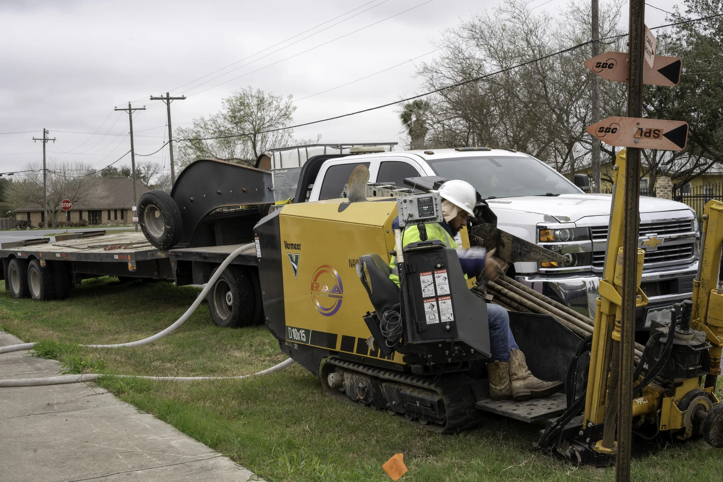 A construction worker operating a small tracked machine on grassy area next to a sidewalk, with a white pickup truck and urban neighborhood in the background.
