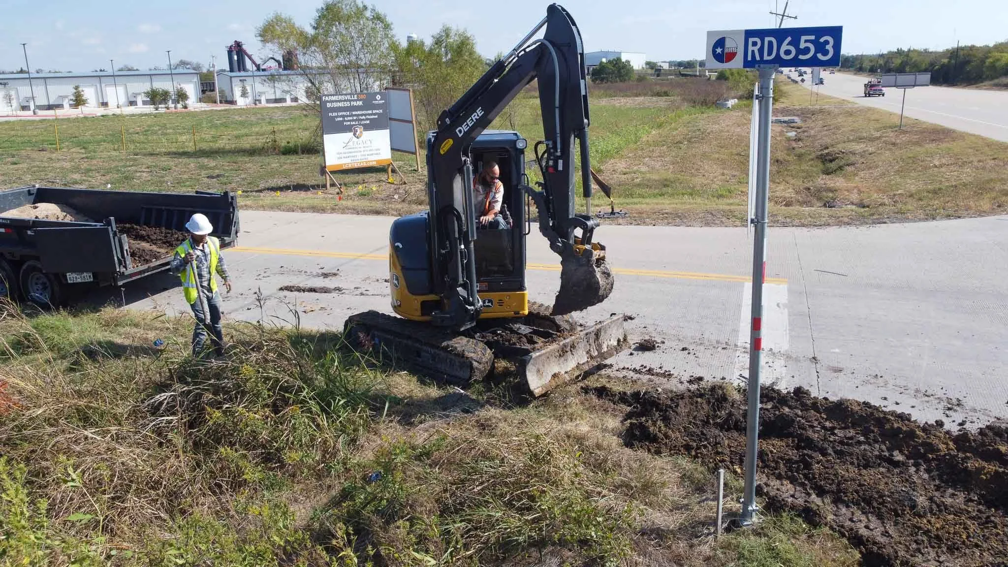 Construction workers operating a small excavator and standing near a trailer on a roadside. The area appears to be under roadwork or maintenance.