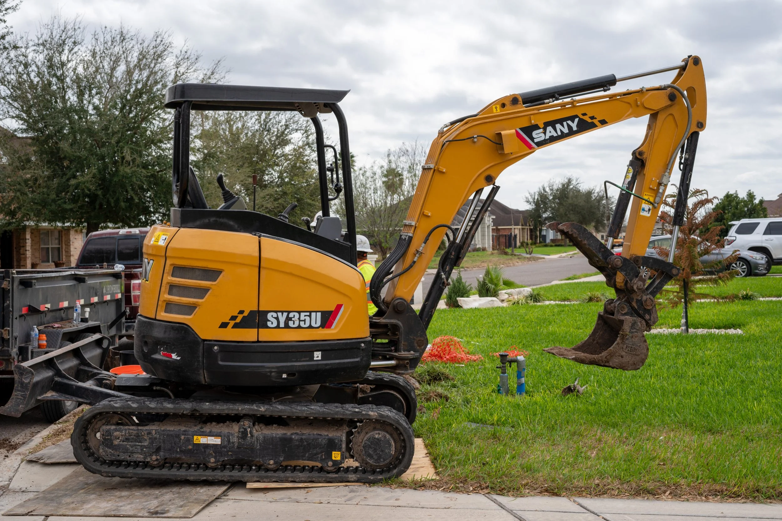 Compact yellow and black excavator with the model number SY35U parked on a grassy area beside a sidewalk in a suburban neighborhood.