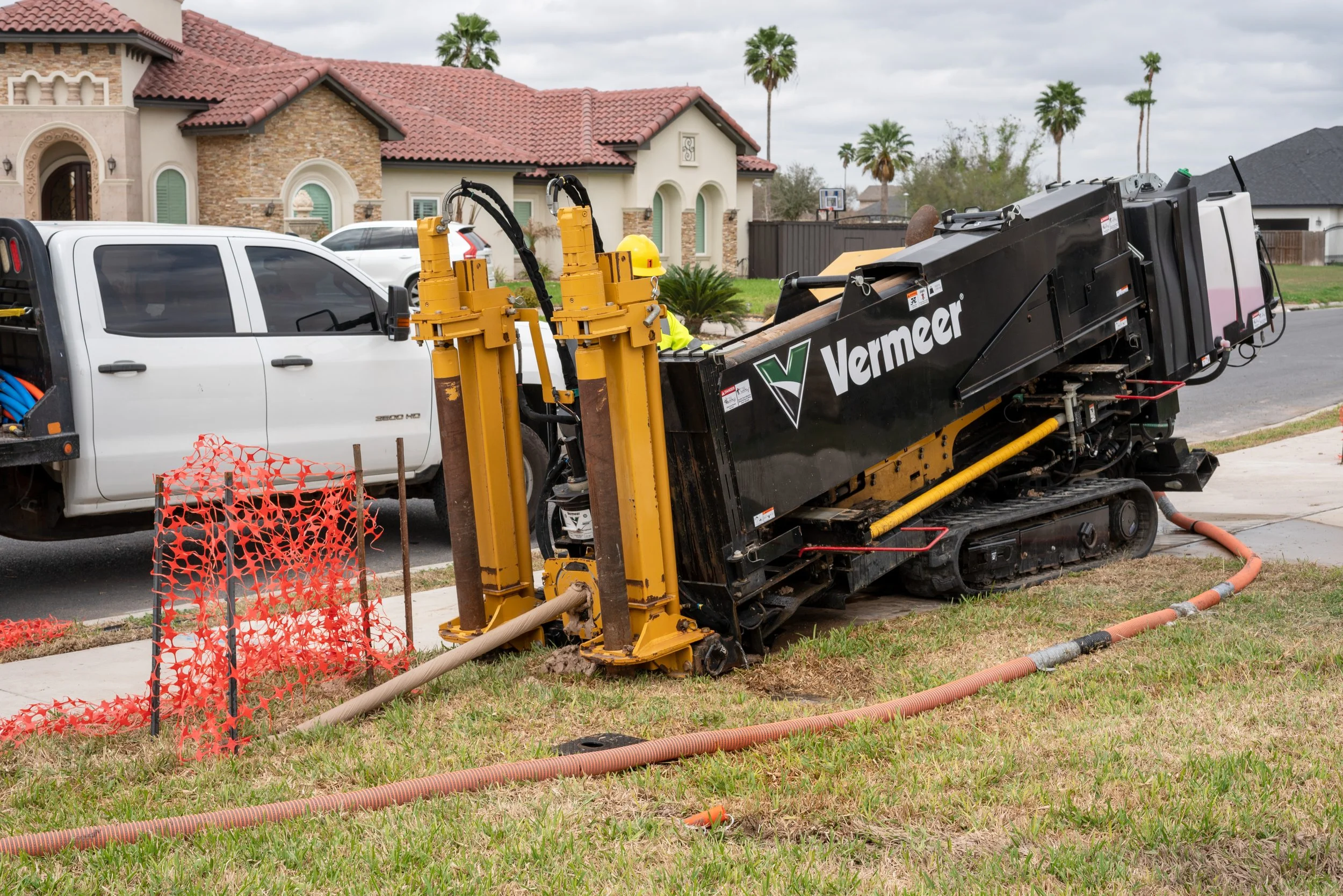 A large Vermeer trenchless drilling machine is working on a residential street, with a worker in safety gear nearby. The machine is positioned on the grass, and a construction barrier is set up around the work area.