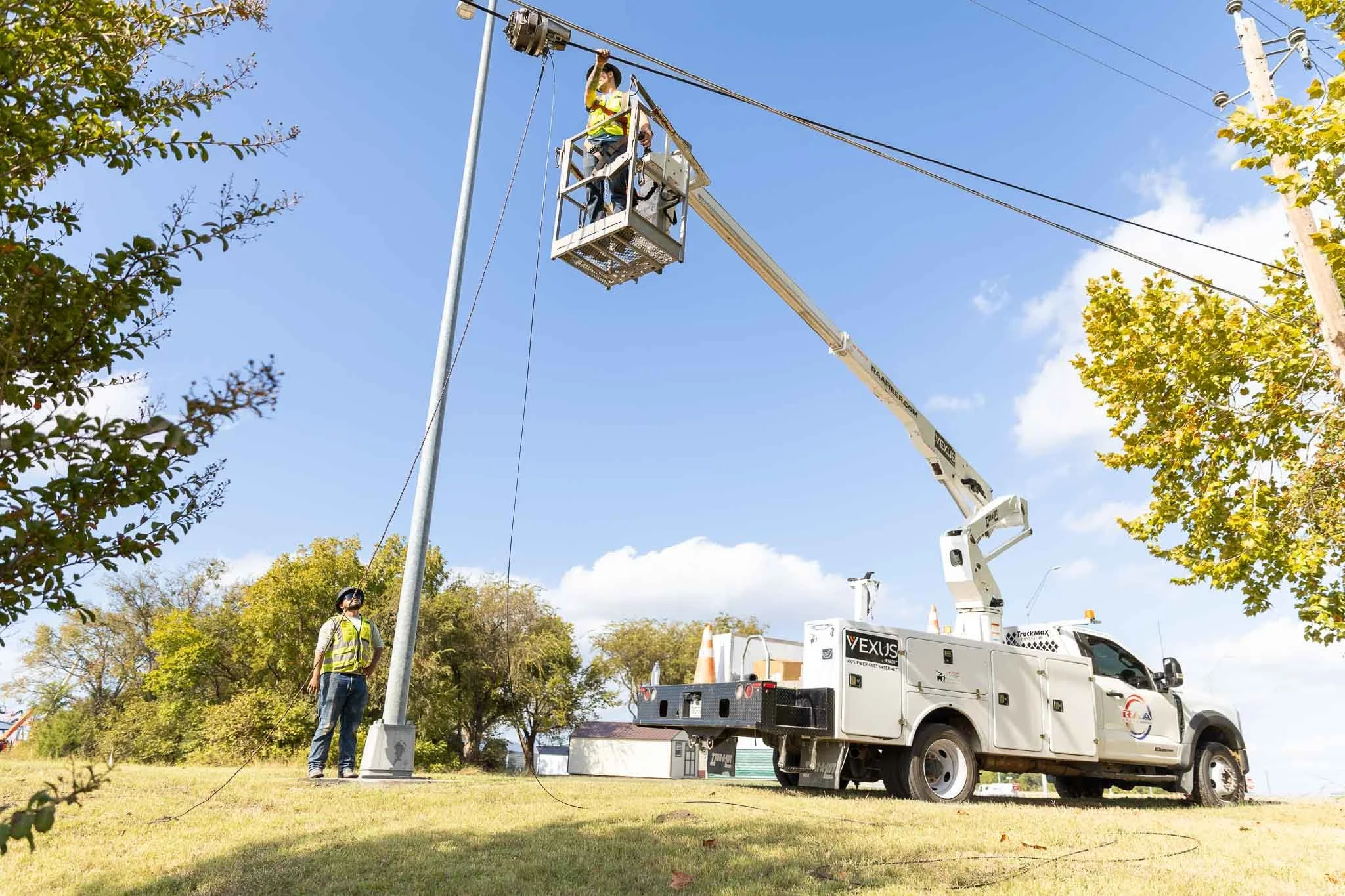 Worker in a bucket lift repairing or inspecting power lines in a park with trees and a blue sky.