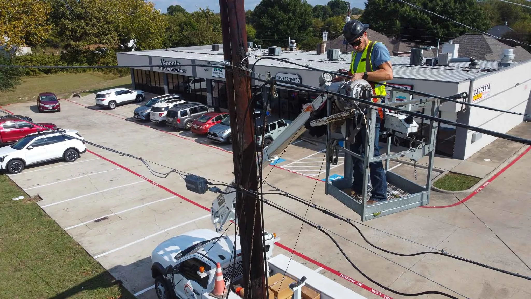 A worker on a crane lift working on utility lines near a parking lot outside a restaurant with multiple parked cars and a building with signs, trees in the background.