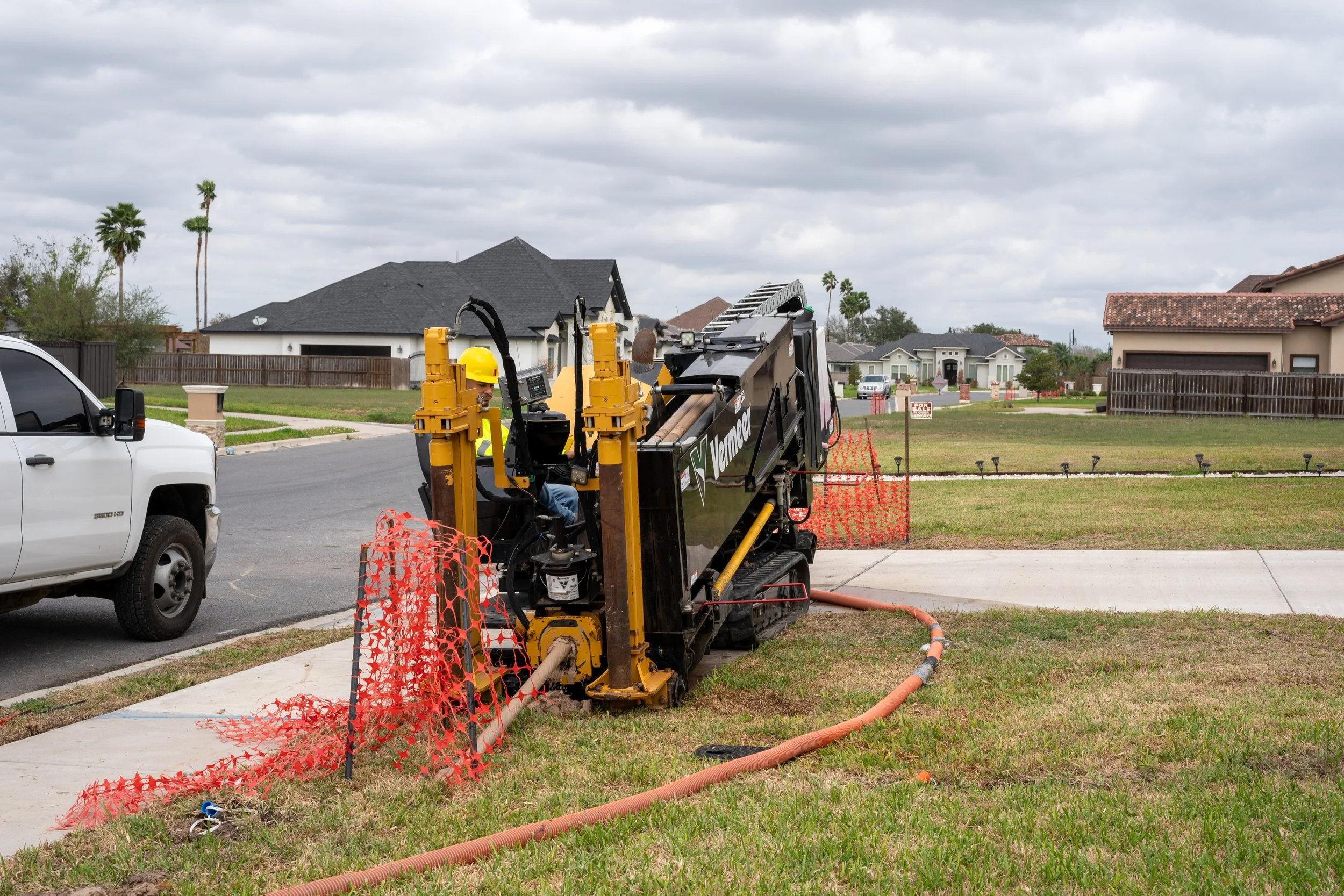 Construction worker operating a trencher on a residential lawn, with a white pickup truck parked nearby and houses in the background.