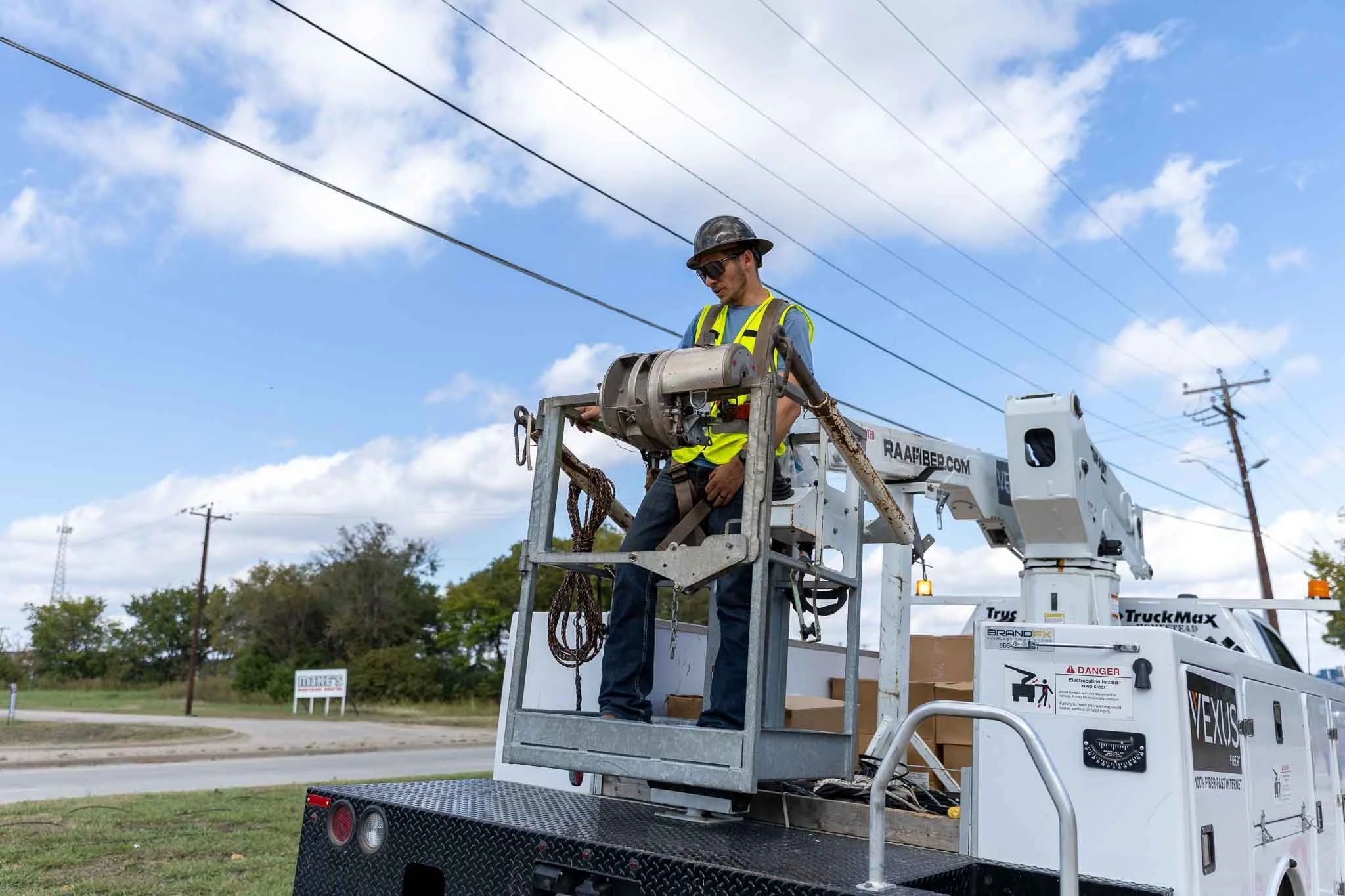 A utility worker wearing a yellow safety vest, gray shirt, jeans, and a helmet working on electrical lines from a utility truck with a cherry picker lift.
