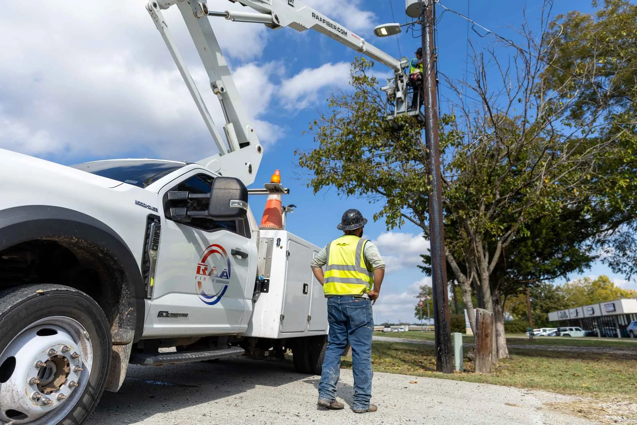 A utility worker in a safety vest and hard hat operating an aerial lift bucket from a bucket truck to work on power lines in a park-like setting with trees and a grassy area.