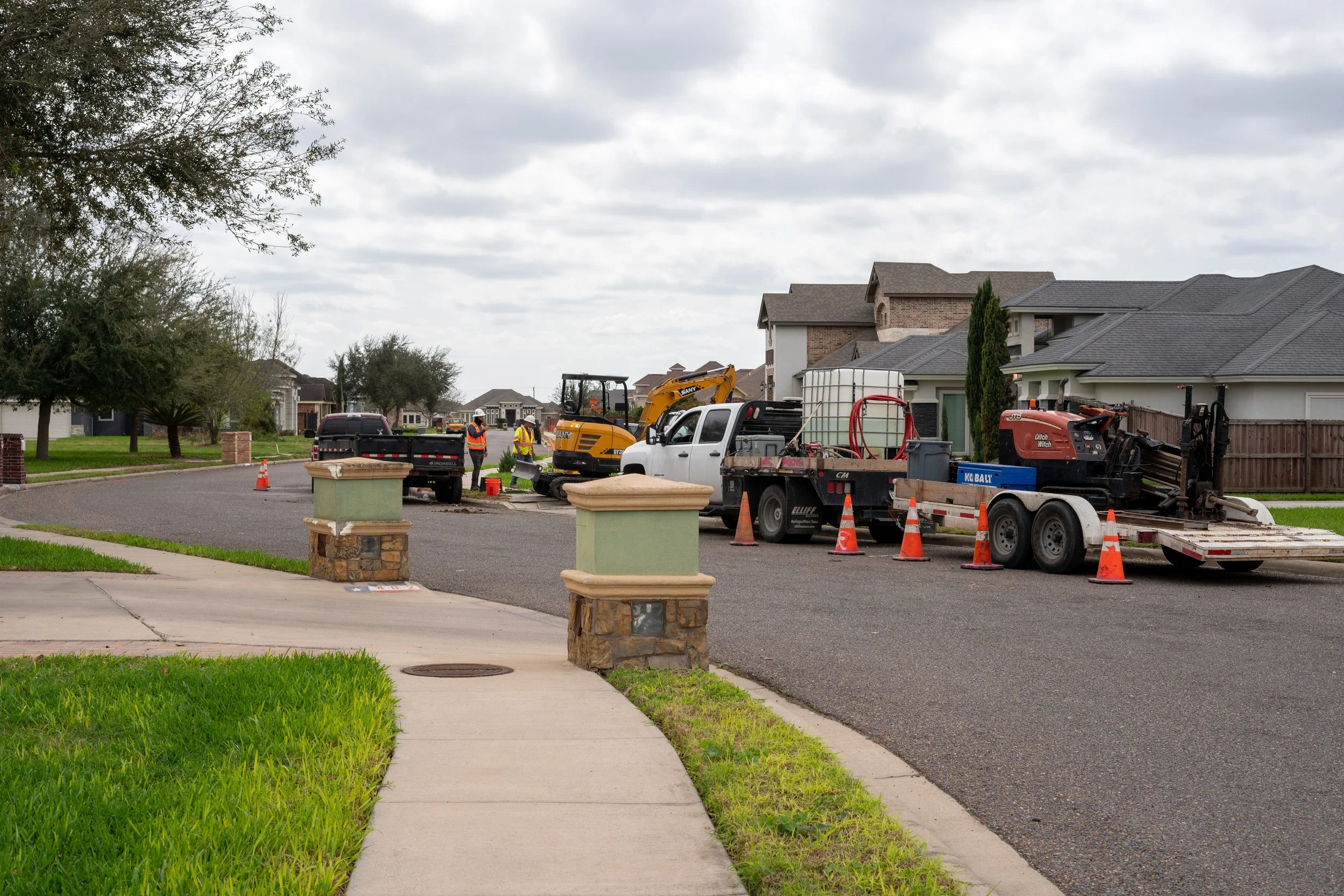 Construction workers working on a residential street with various construction vehicles and equipment, orange cones, and houses in the background.