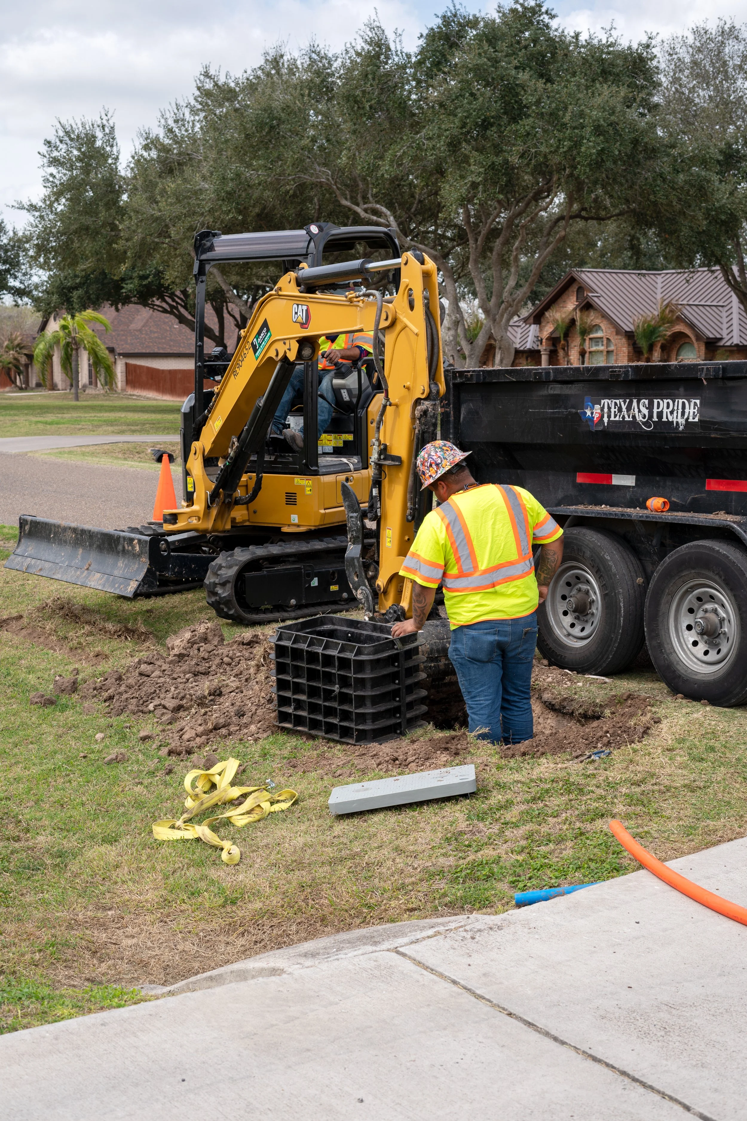 Worker in a yellow safety vest and floral hard hat installing a fiber optic line in a residential neighborhood using a small excavator, with a black Texas Pipe truck nearby and orange traffic cones on a cloudy day.
