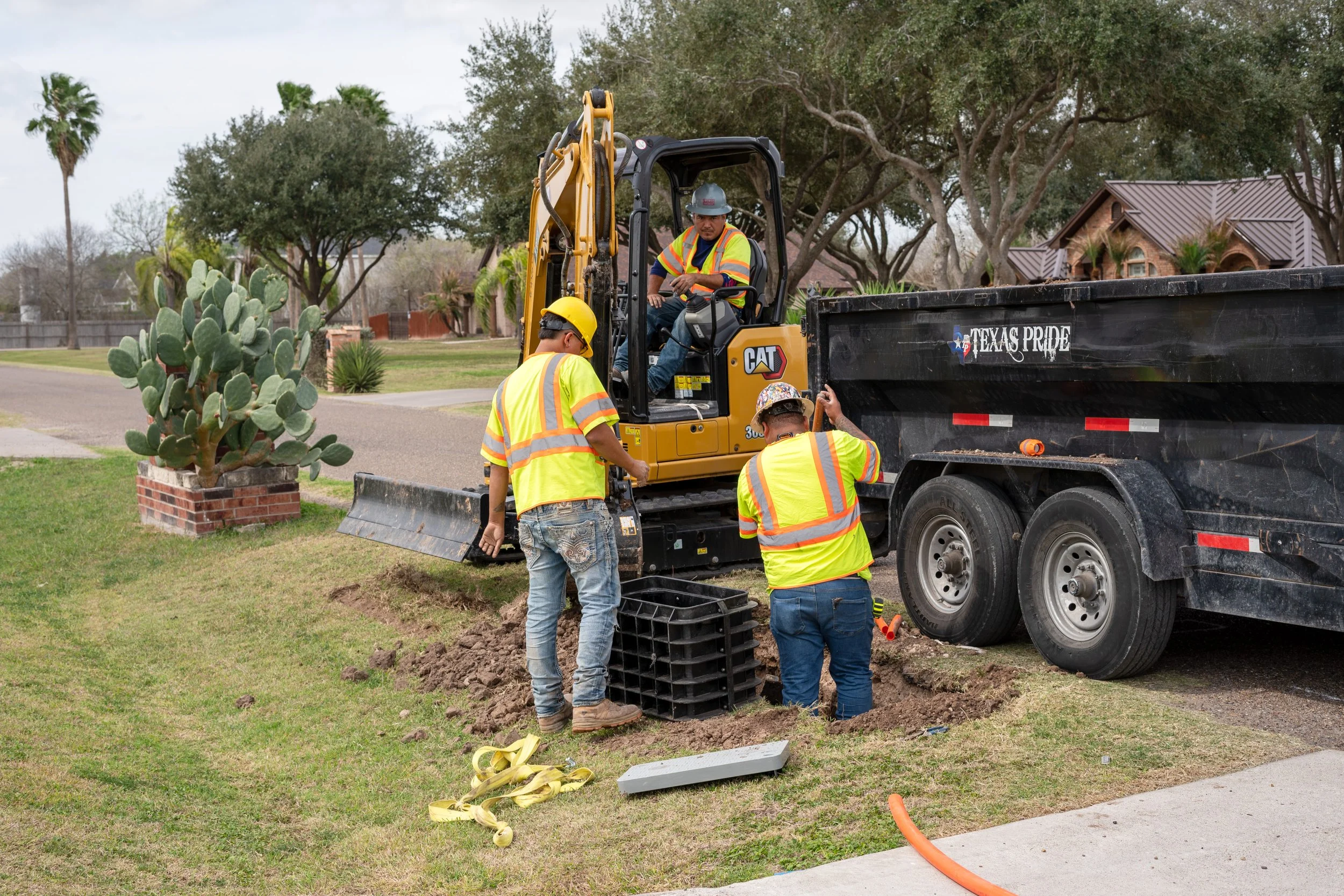 Construction workers digging a trench on a grassy area beside a road, using a small excavator, with a truck labeled "Texas Pride" nearby.