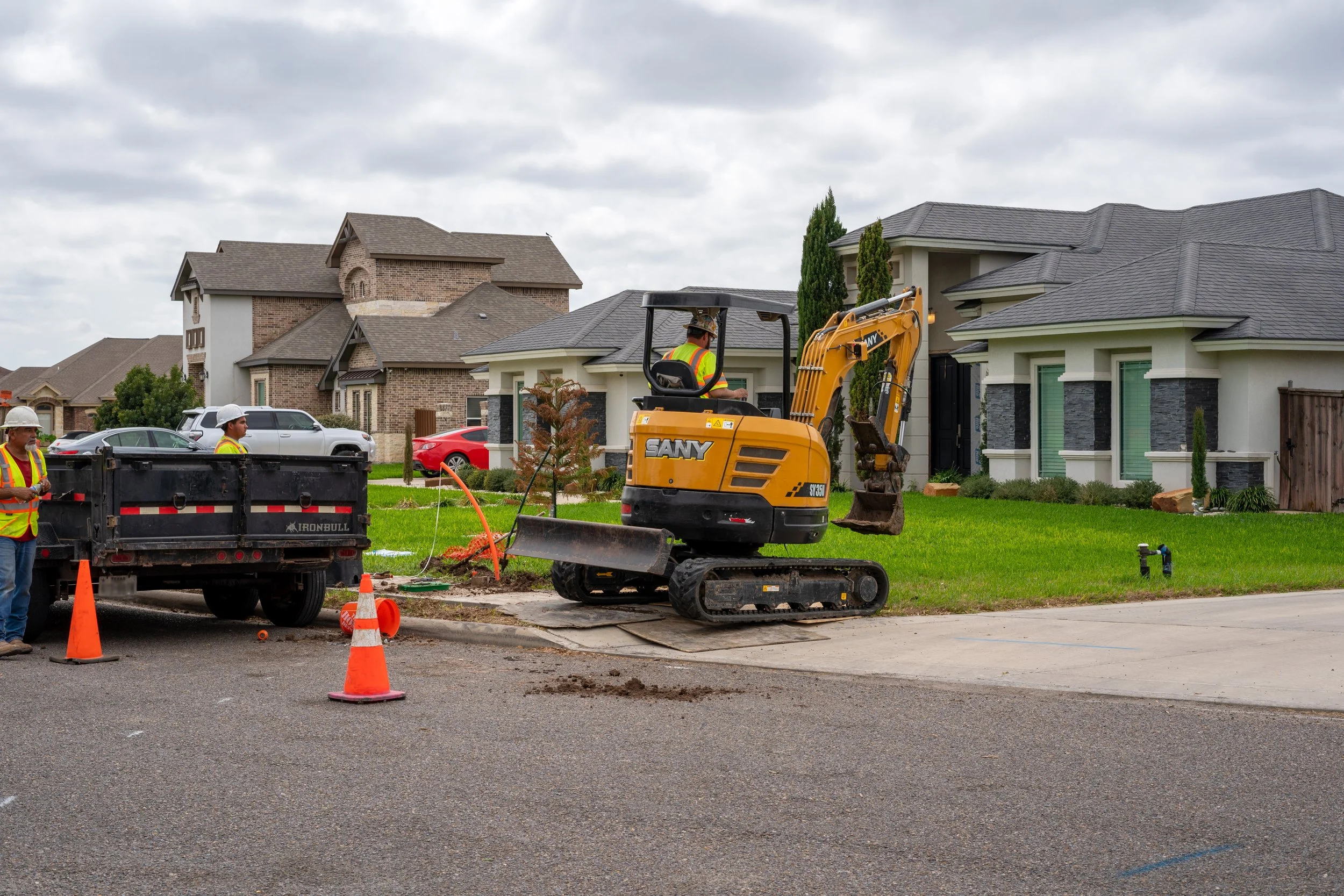 Construction workers are working on a residential street, using a small yellow excavator to dig near a curb in front of a house, with safety cones and a truck nearby.