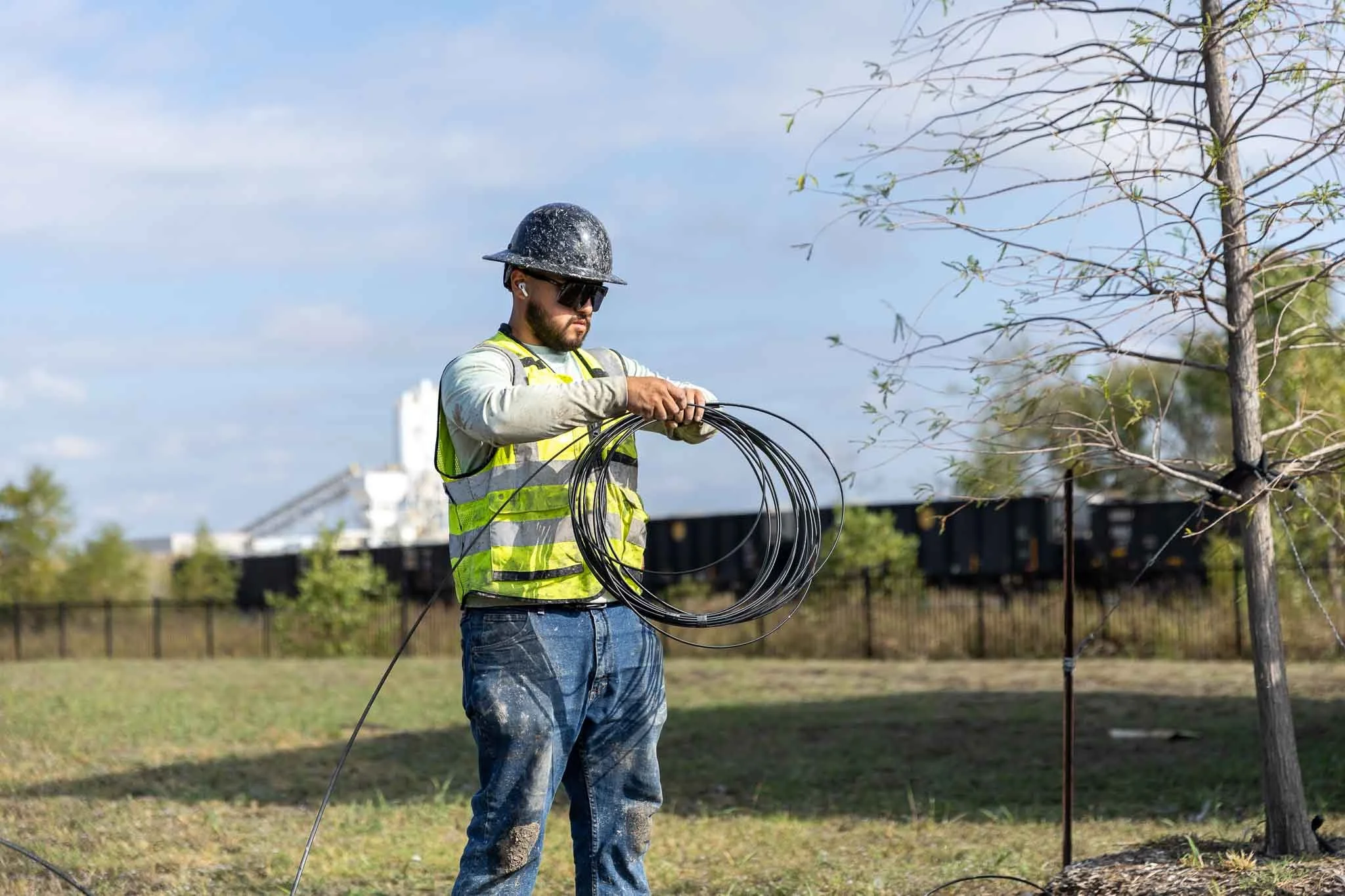 A man wearing a helmet, sunglasses, and a high-visibility safety vest handling black electrical wiring outdoors near a young tree with undergrowth, with a fence and train tracks in the background.