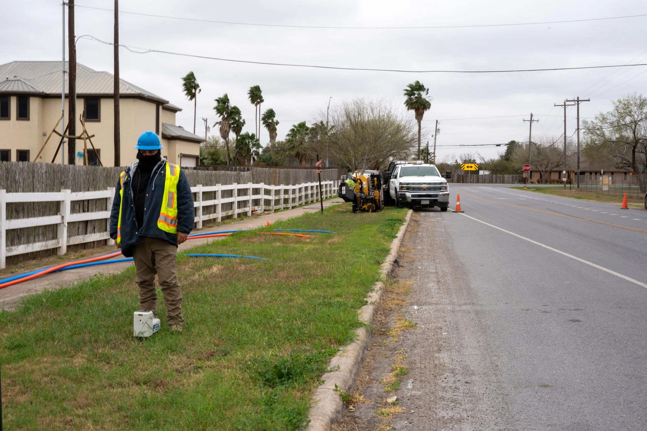 A utility worker wearing a blue hard hat and a safety vest standing on the grass near a sidewalk, with work equipment nearby. There is a white pickup truck and a work truck parked on the side of the road, which is marked off with orange traffic cones
