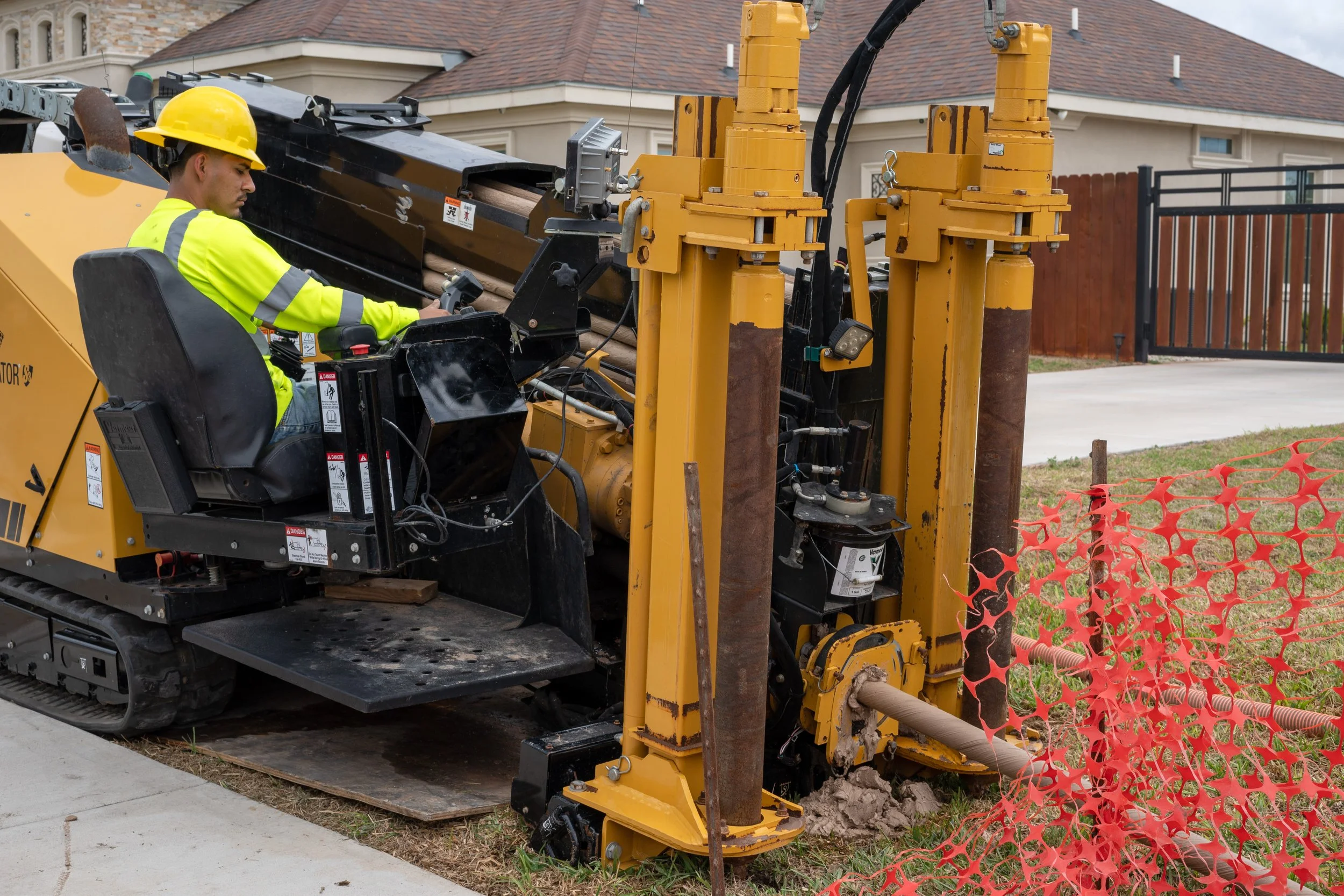 A construction worker in a yellow safety vest and helmet operating a trenching machine on a sidewalk next to a house, with orange safety fencing nearby.