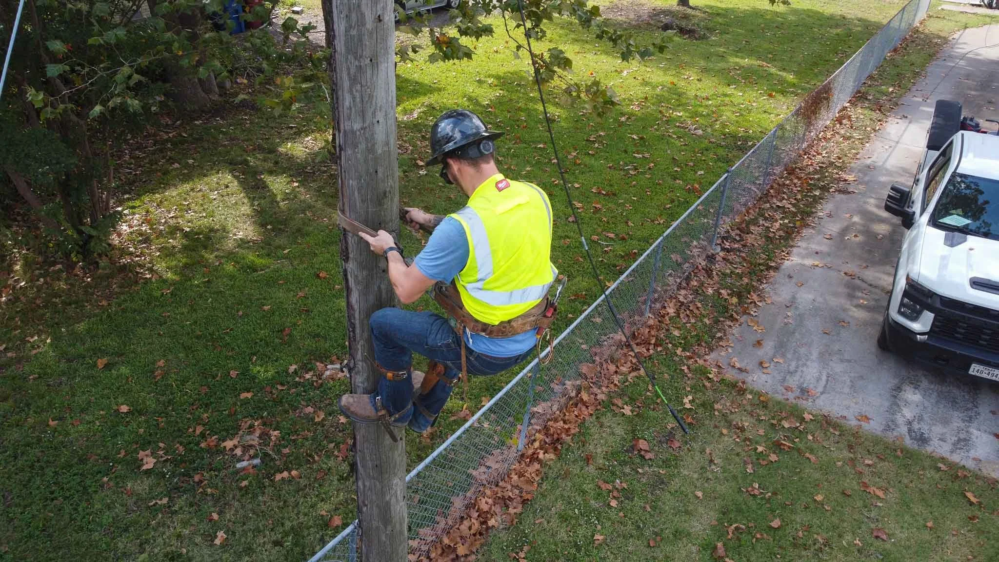 A worker wearing a safety helmet, a reflective yellow vest, and a harness is sitting on a wooden utility pole, working on electrical wires. The worker is holding tools and is positioned above a fenced grassy area next to a parked white truck on a con