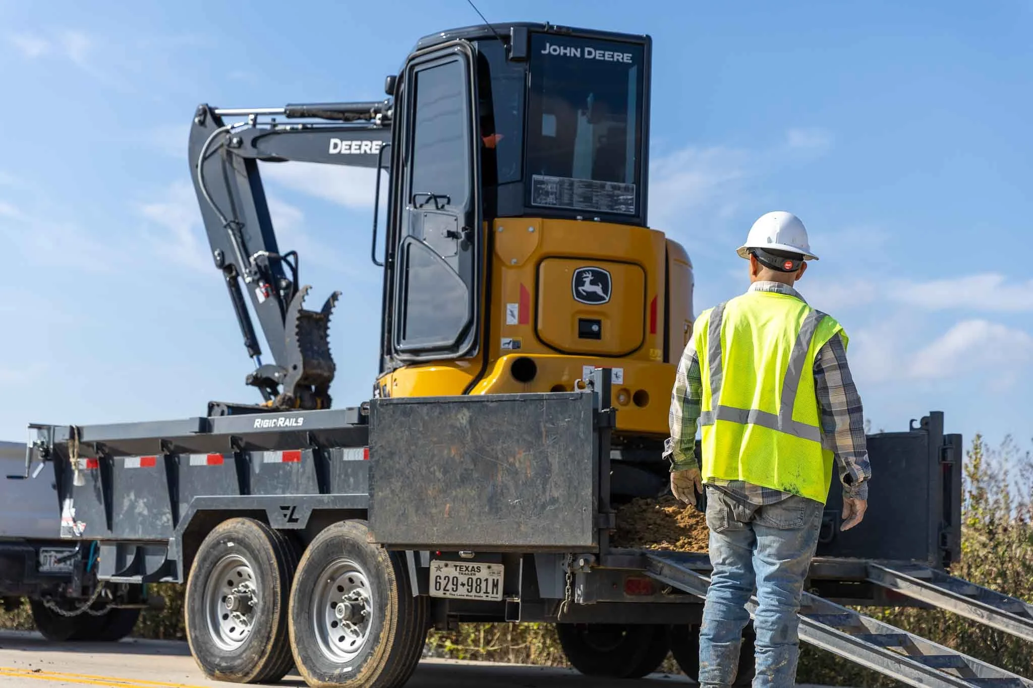 Construction worker in a yellow safety vest and white helmet observing a small John Deere excavator on a flatbed trailer. The trailer is parked on a road under a partly cloudy sky.