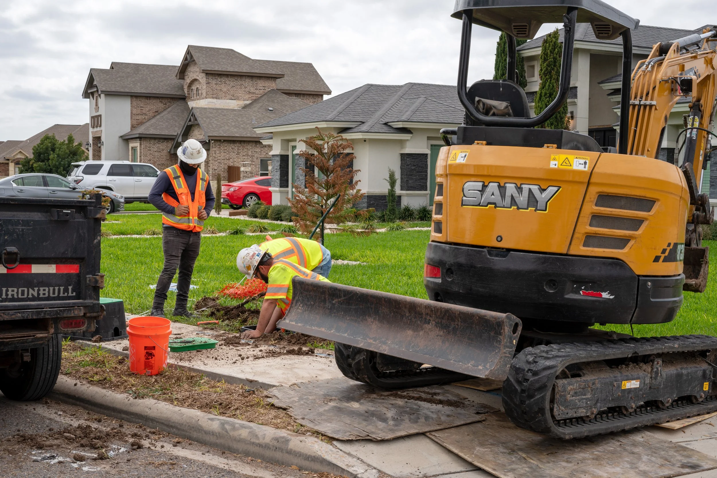 Two construction workers in safety vests and helmets working on a sidewalk, with a small excavator nearby, in a residential neighborhood.