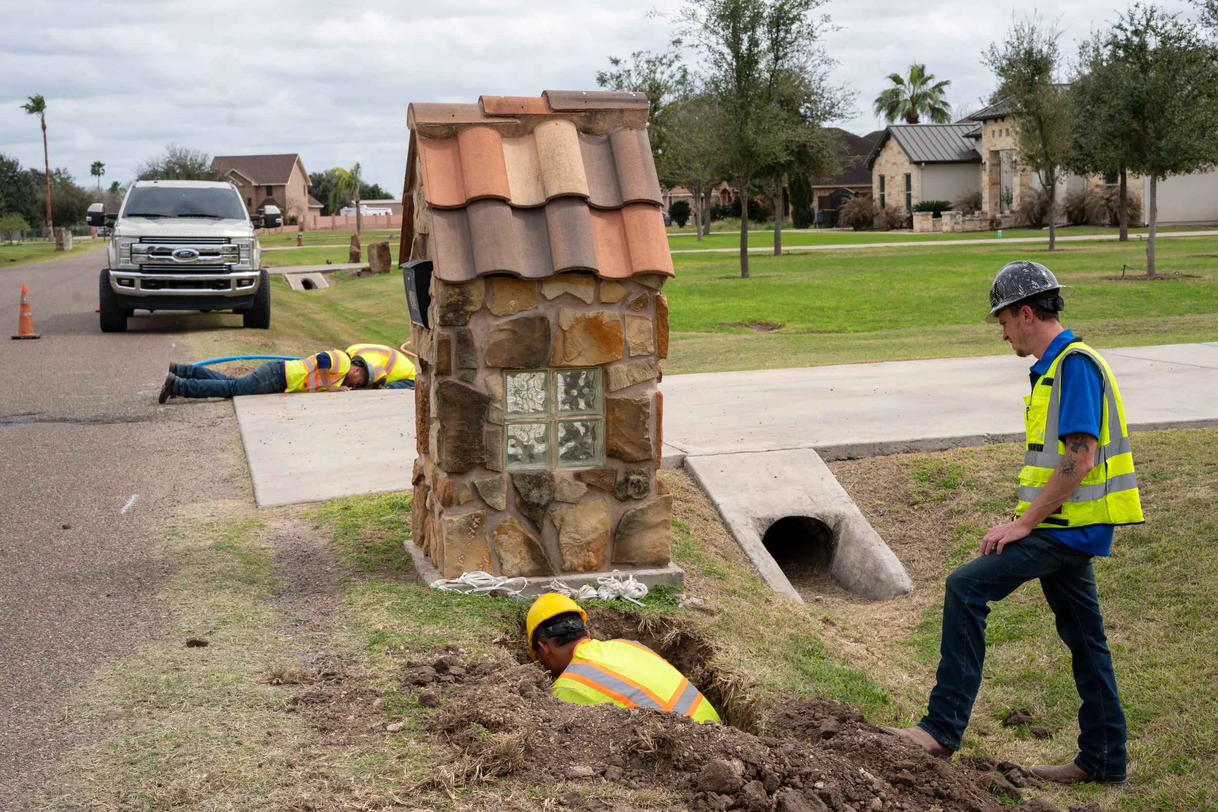 Construction workers installing utility lines near a stone and brick mailbox on a residential street. One worker is inside a hole, another is lying on the sidewalk, and a third is standing nearby watching.