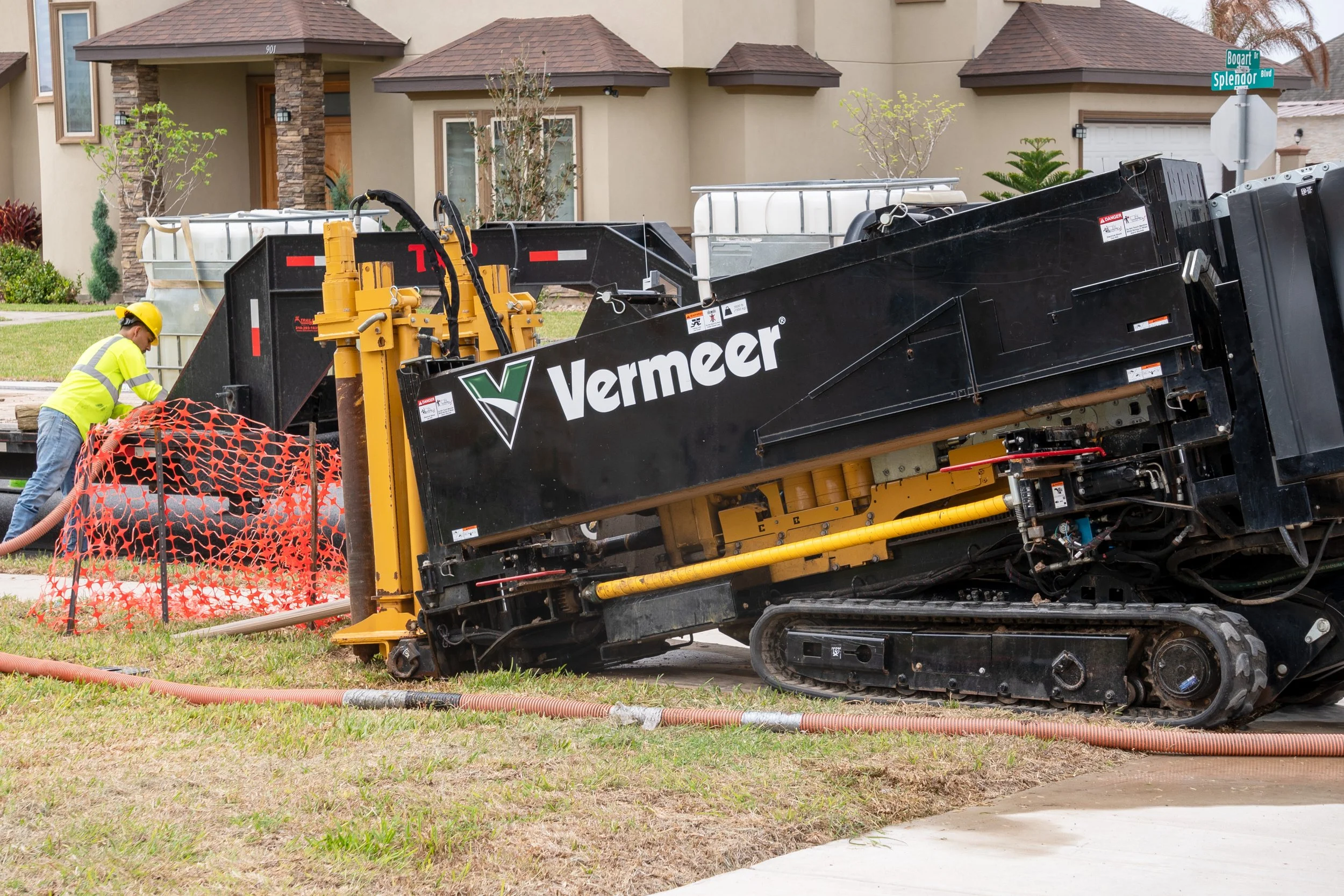 A worker in a yellow safety shirt and helmet operating a large Vermeer trenchless drilling machine on a residential street.