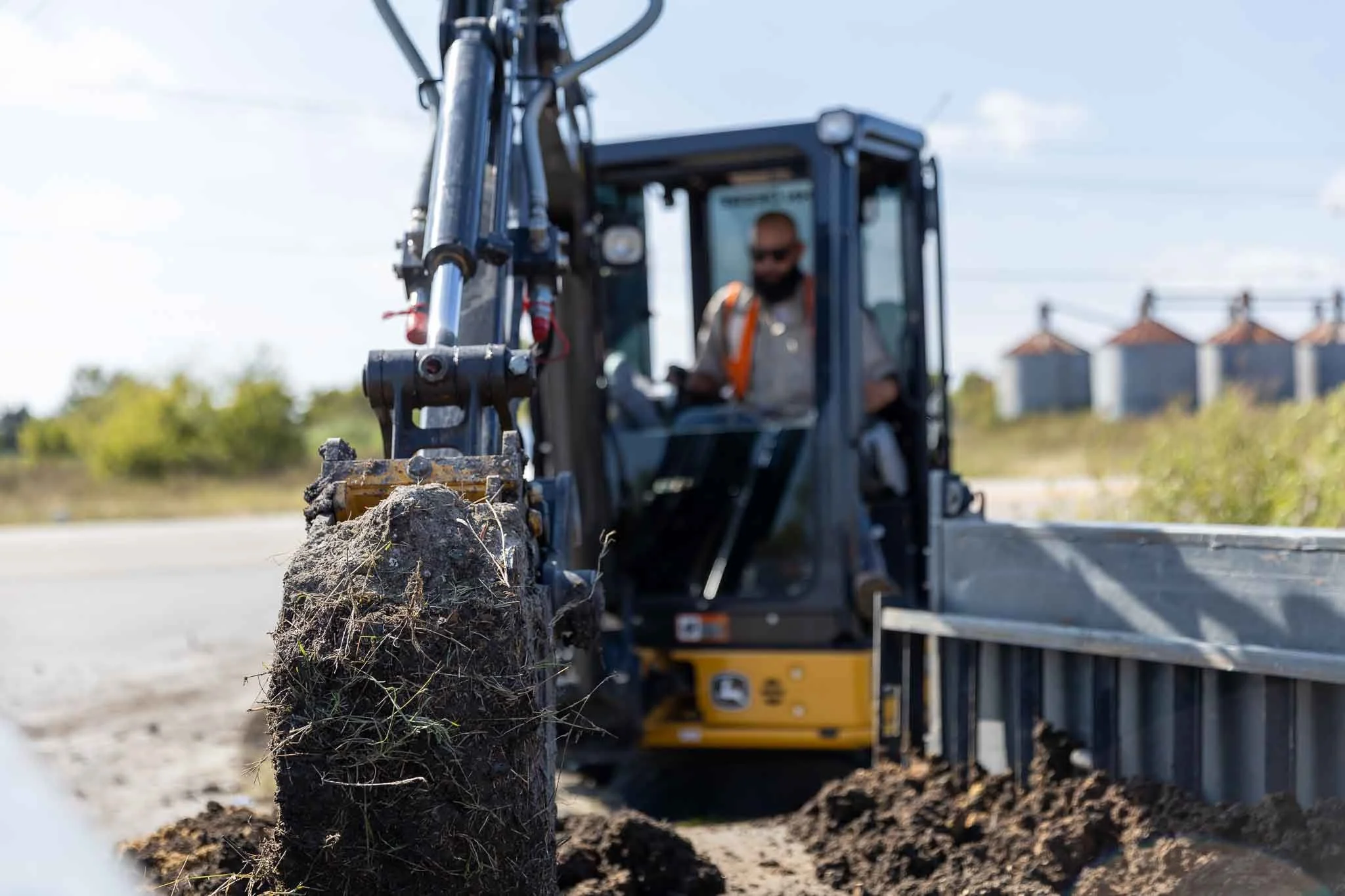 A construction worker operating a small excavator on a dirt construction site with silos in the background.