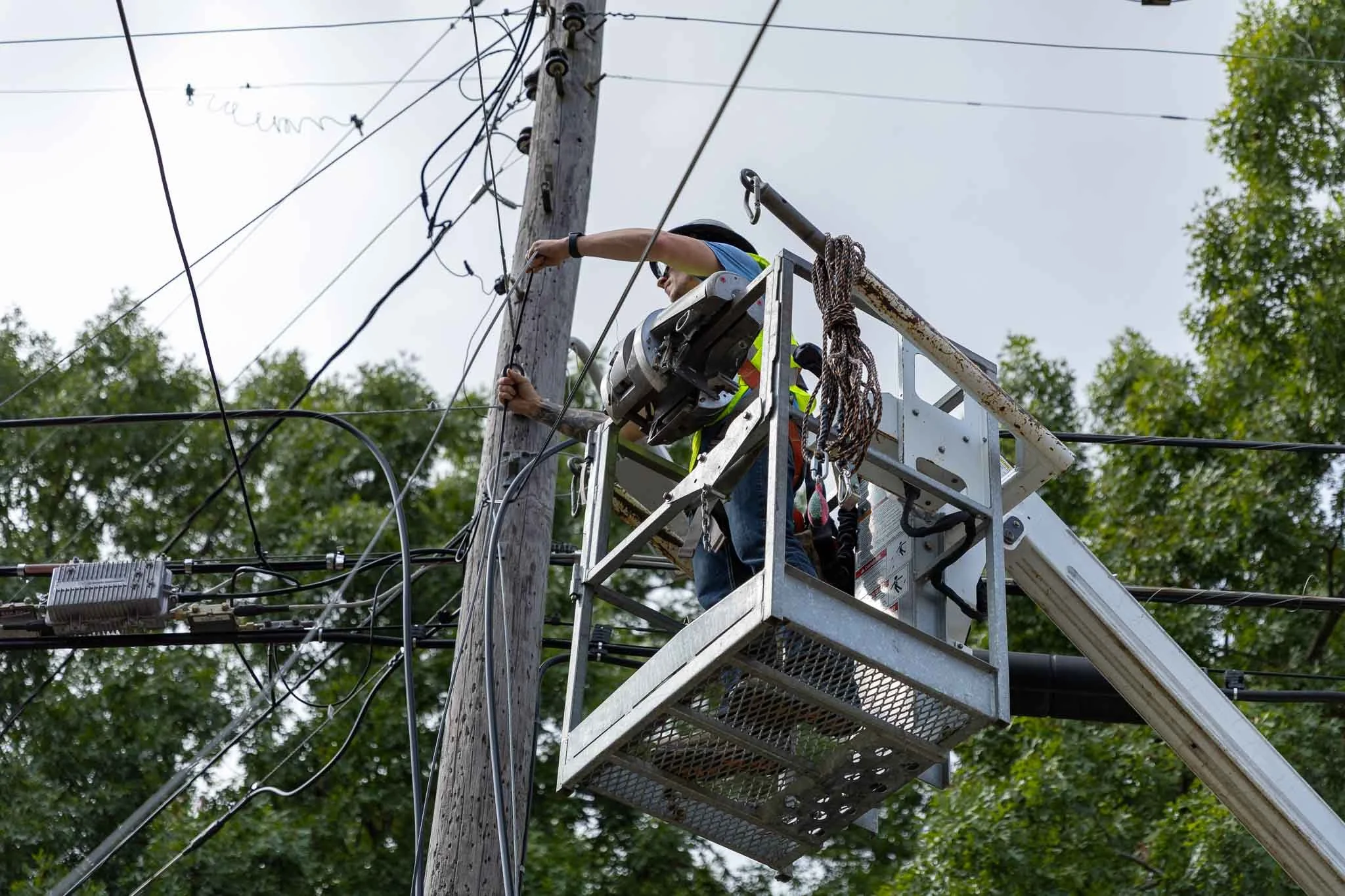 A utility worker in a safety vest and helmet working on electrical wires on a utility pole from a raised bucket lift.