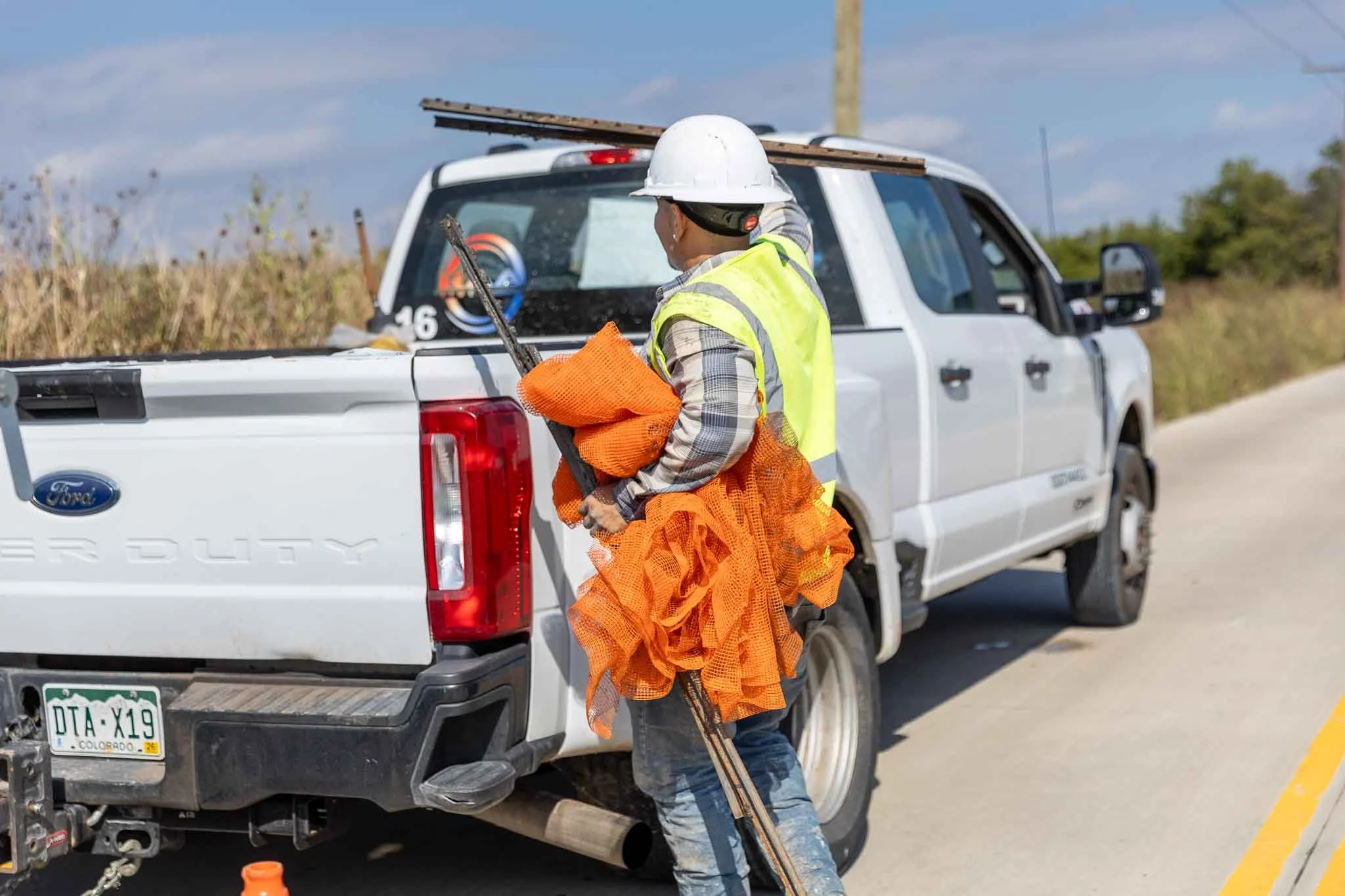 Worker wearing a white safety helmet and yellow reflective vest standing next to a white pickup truck on a rural road, holding orange safety vests and metal poles.