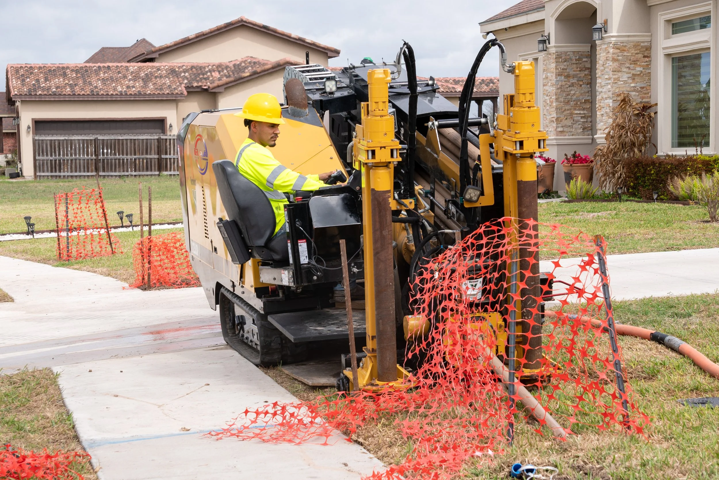 A construction worker in a yellow safety helmet and high-visibility jacket operates a small tracked drilling machine on a residential lawn, with orange safety fencing around the work area.