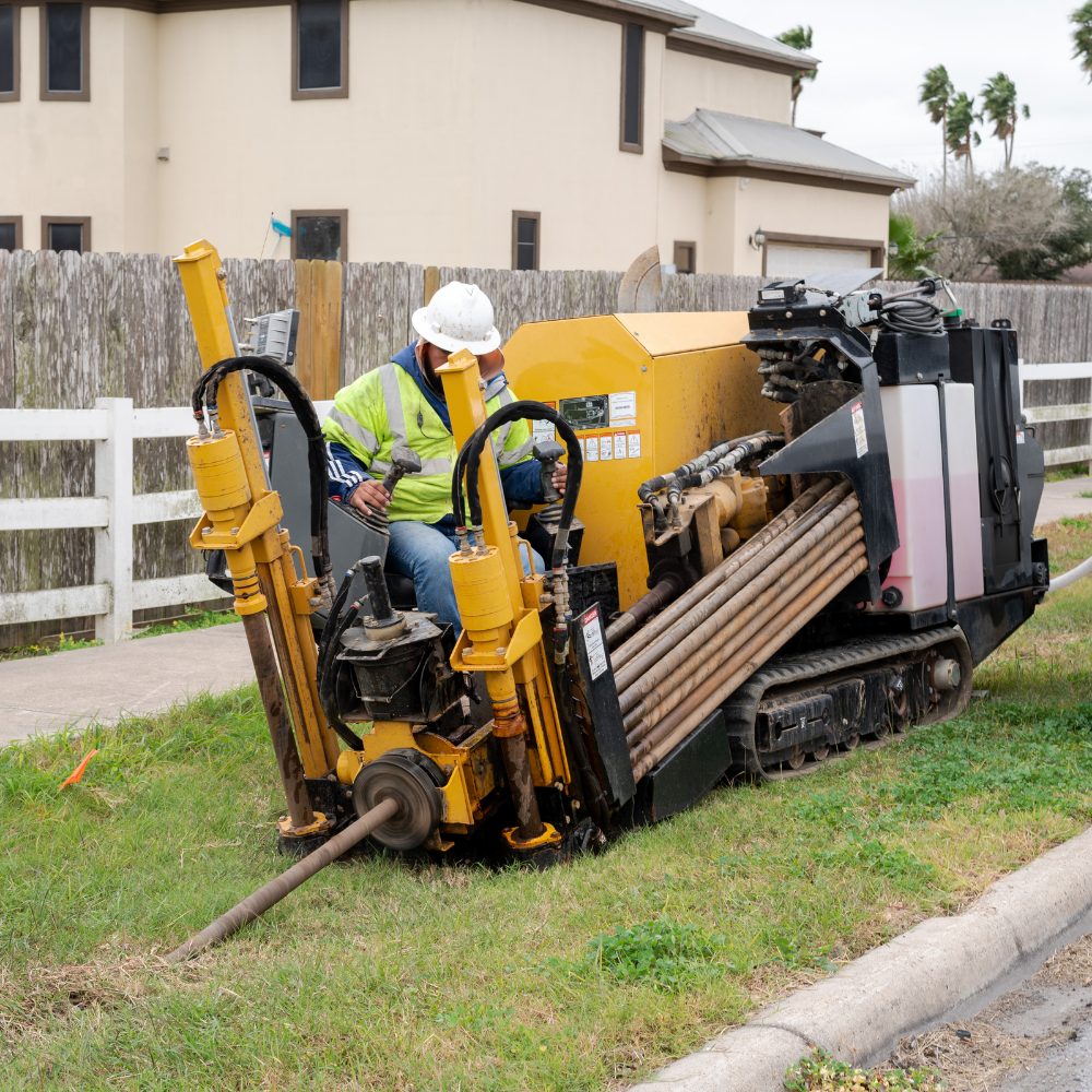 Worker operating a road drilling machine on a lawn near a residential area with a white fence and a house in the background.